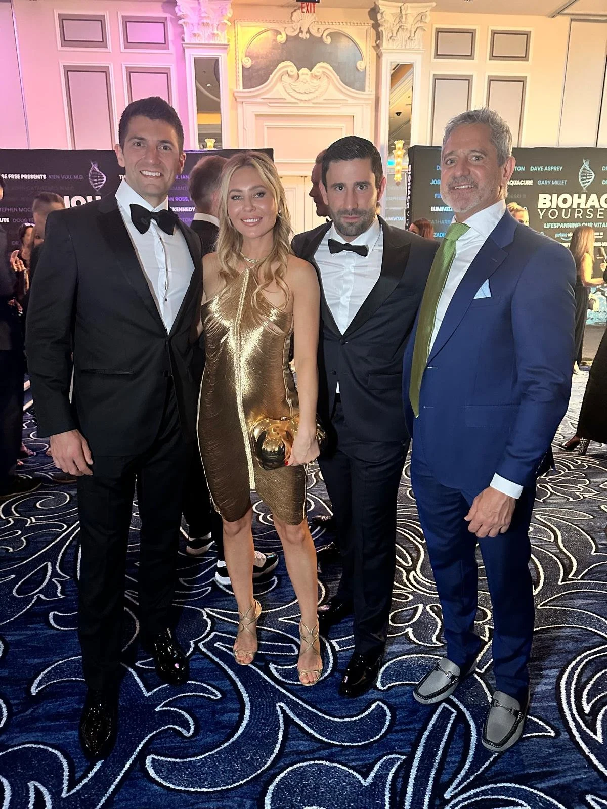 Four people dressed in formal attire at an event, with two men in black tuxedos, a woman in a gold dress, and another man in a blue suit with a green tie. They are standing on a patterned carpet with a decorated backdrop.