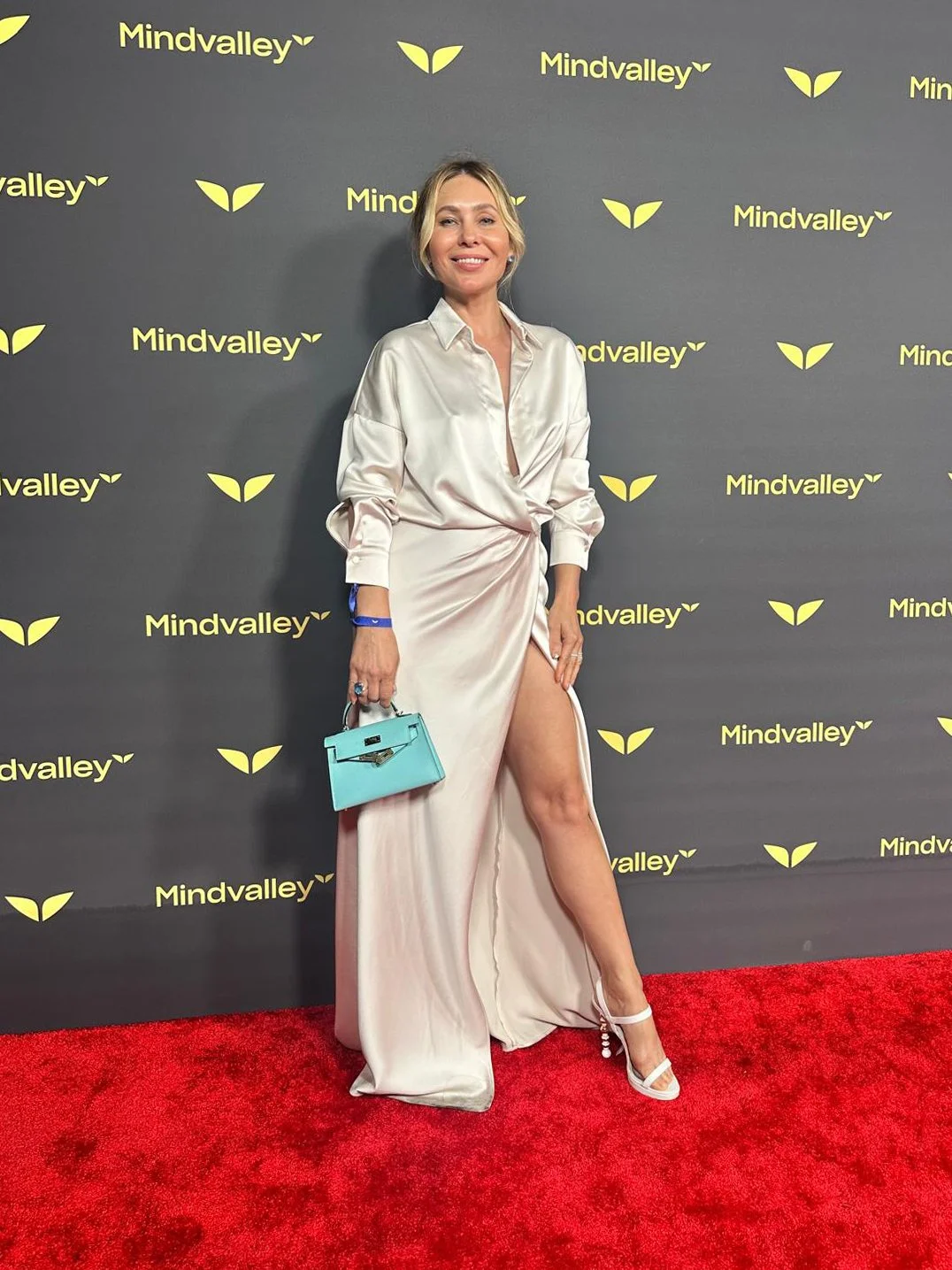 A woman in a shiny, cream-colored dress with a high slit, standing on a red carpet against a backdrop with the 'Mindvalley' logo and yellow leaves. She is holding a small, turquoise handbag and smiling at the camera.