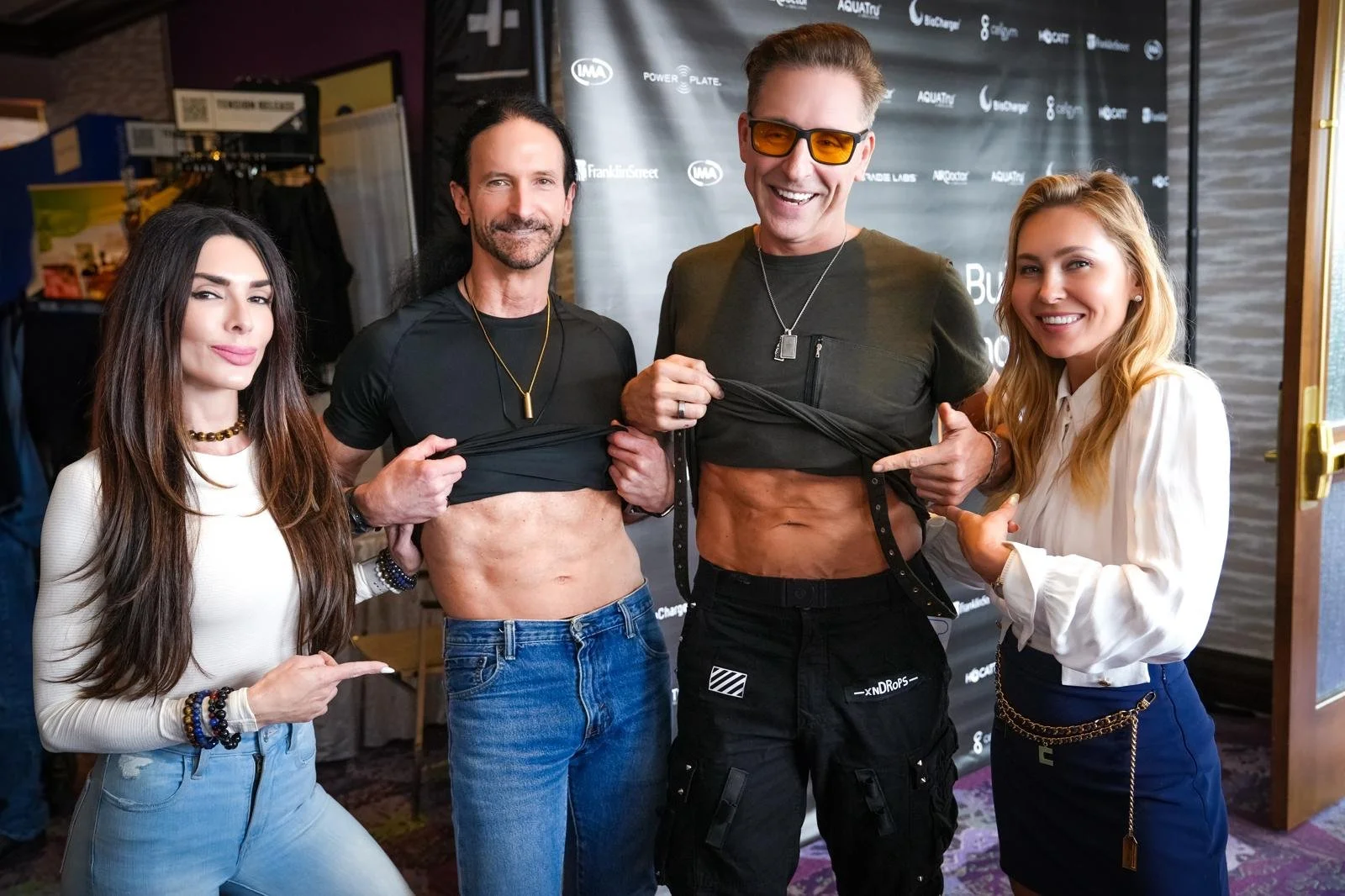 Four people standing together at an event, lifting their shirts to reveal their abs, with two pointing and smiling at the camera.