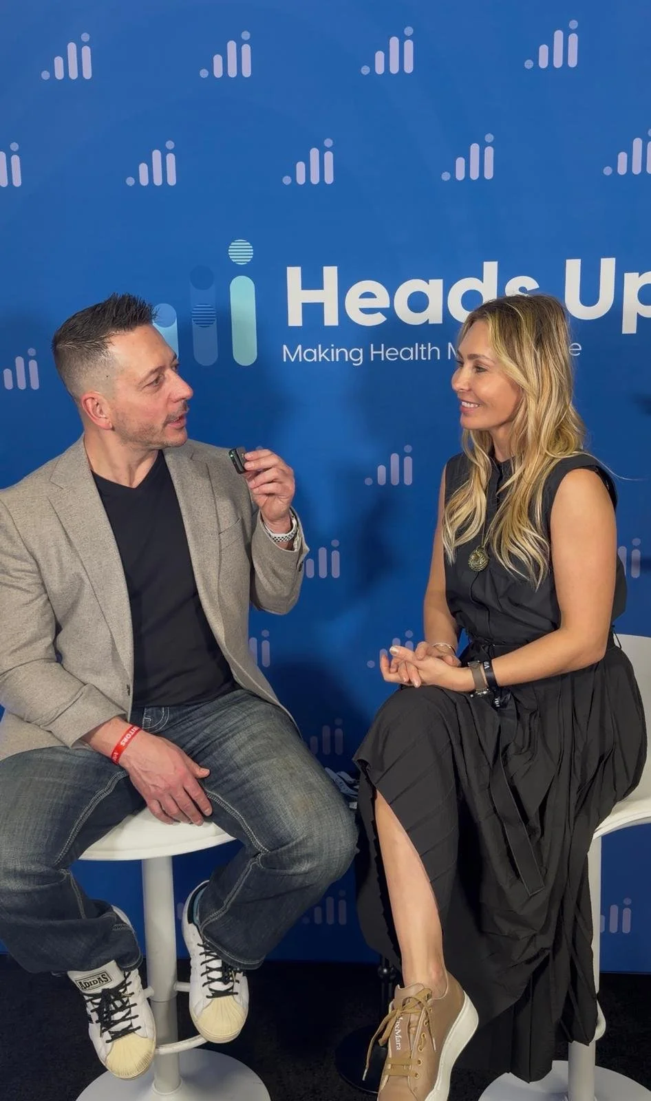 A man and woman having a conversation during an interview at a health conference, with a blue backdrop featuring the 'Heads Up' logo and slogan 'Making Health Made Easy'.