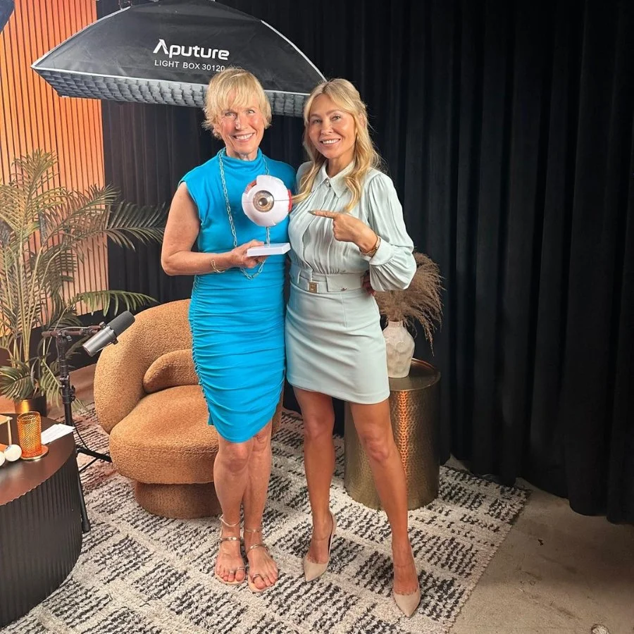 Two women standing indoors, one holding an award and the other pointing at it, under a large studio light, with a beige chair, plants, and decorative items around them.