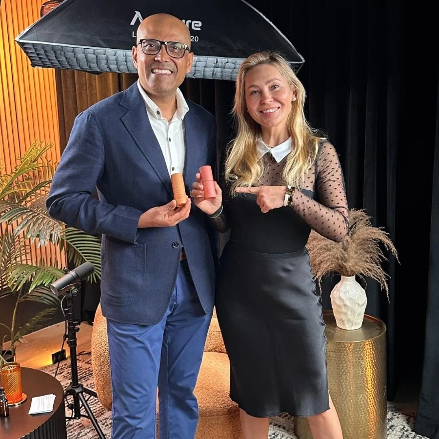 Two people, a man and a woman, standing indoors, smiling, holding lipstick products, with photography equipment and a large umbrella behind them, and a decorated table with a potted plant and a vase nearby.