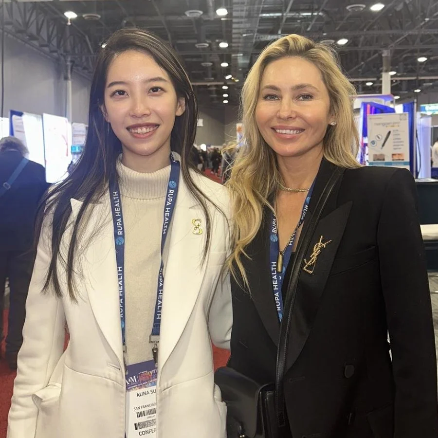 Two women at a conference, both wearing conference badges, smiling and standing close together in a large indoor event space.