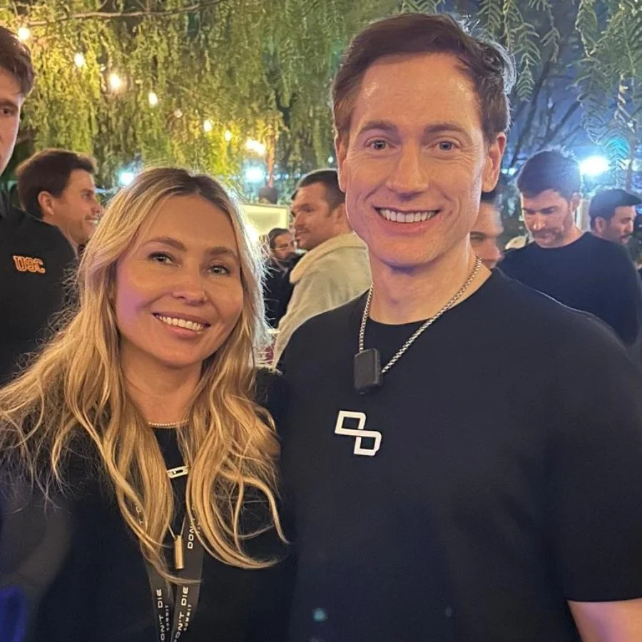 A smiling woman with blonde hair and a man with short brown hair standing together at an outdoor event at night, with other people and string lights in the background.