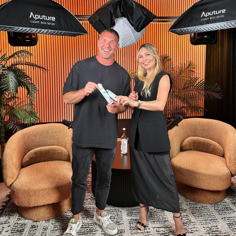 Two smiling people, a man and a woman, stand in a professional photography studio with orange vertical panel wall, surrounded by studio equipment and plants. The man is holding a small tube, and the woman is touching it. There are two modern beige so