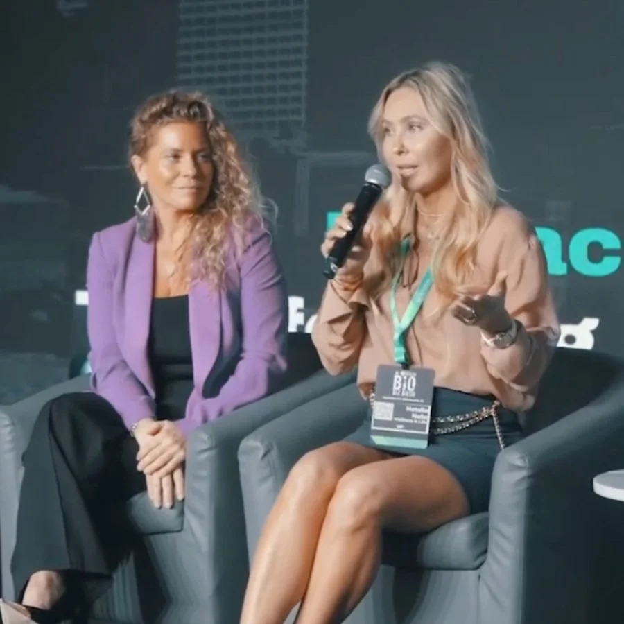 Two women sitting on stage, one speaking into a microphone and the other listening, at a conference or panel discussion.