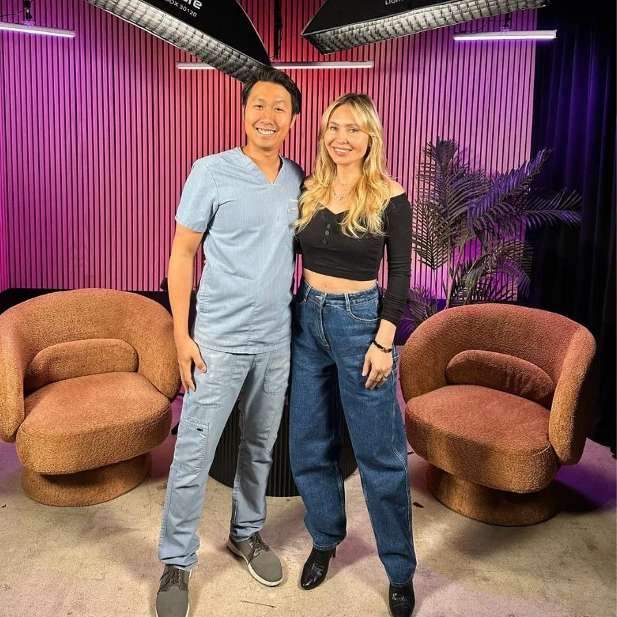 Two people, a man and a woman, smiling and posing for a photo in a colorful studio with pink and purple lighting, chairs, and plants around them.