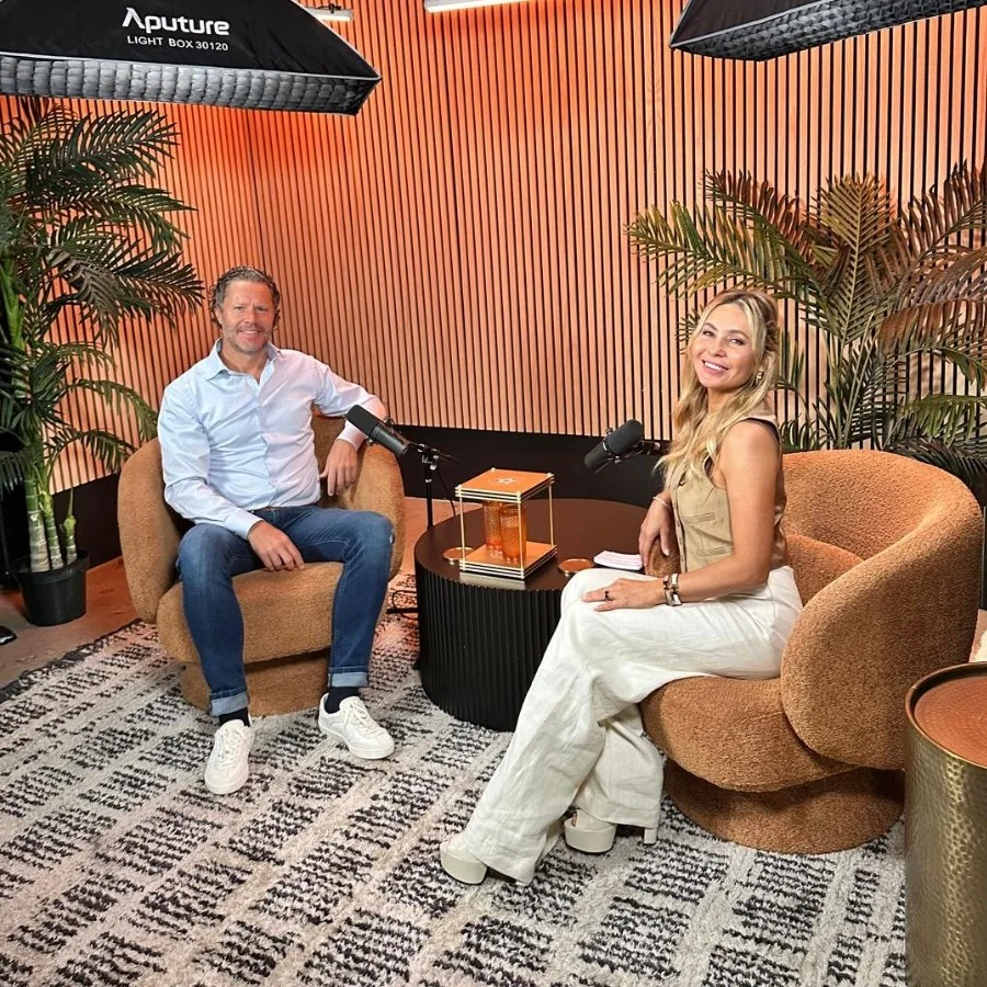 Two people sit in a podcast recording studio with orange walls and large plants. The man on the left is wearing a light blue shirt and jeans, smiling. The woman on the right has long blonde hair and is wearing a tan top and white pants, also smiling.