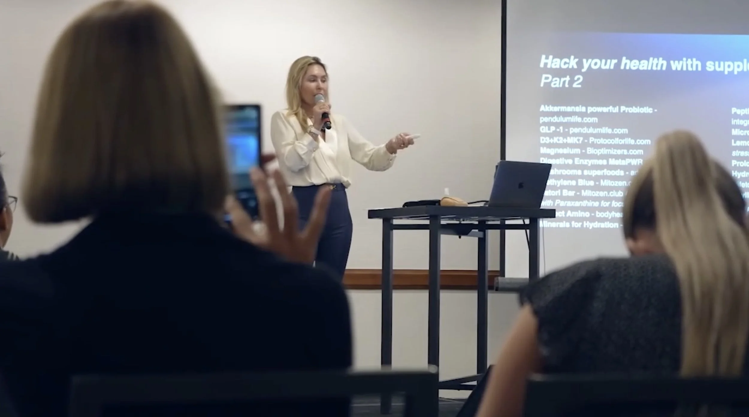 A woman giving a presentation in a conference room with an audience taking pictures, with a slide titled 'Hack your health with supplements'.