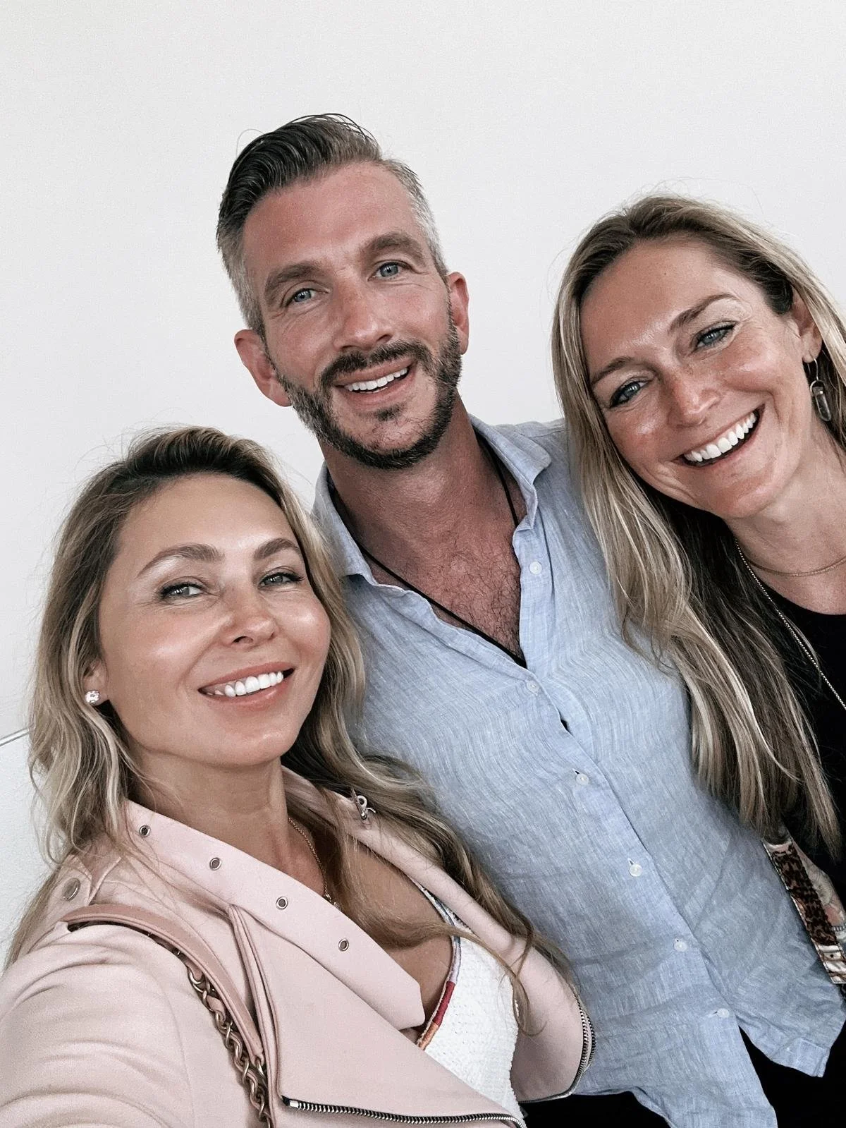 Three smiling adults, two women and one man, taking a selfie against a plain white wall.