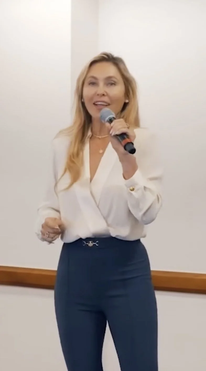 A woman with long blonde hair holding a microphone, speaking or singing in front of a plain white wall.