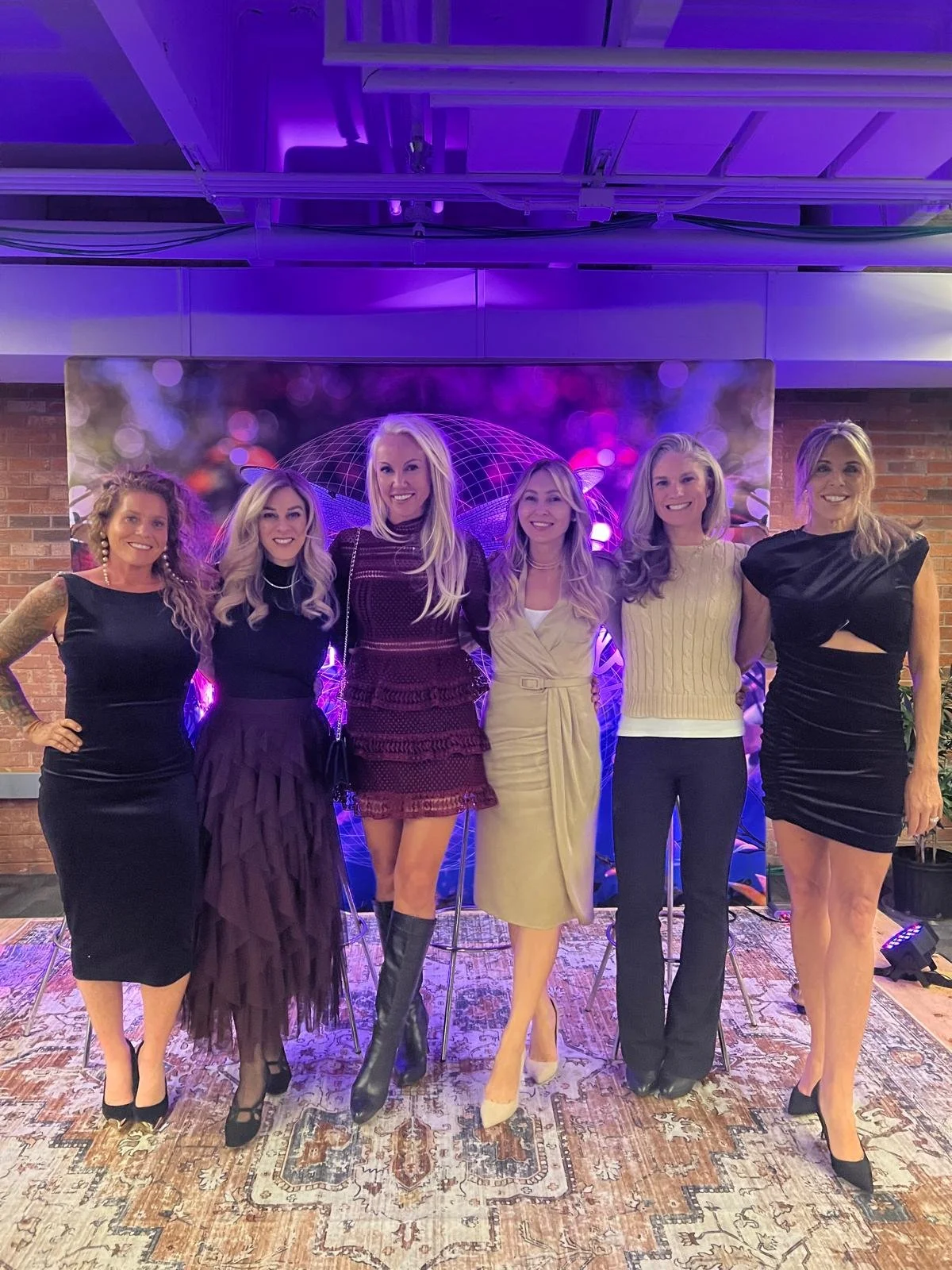 Six women dressed in formal and semi-formal attire standing together indoors in front of a colorful backdrop with purple lighting.
