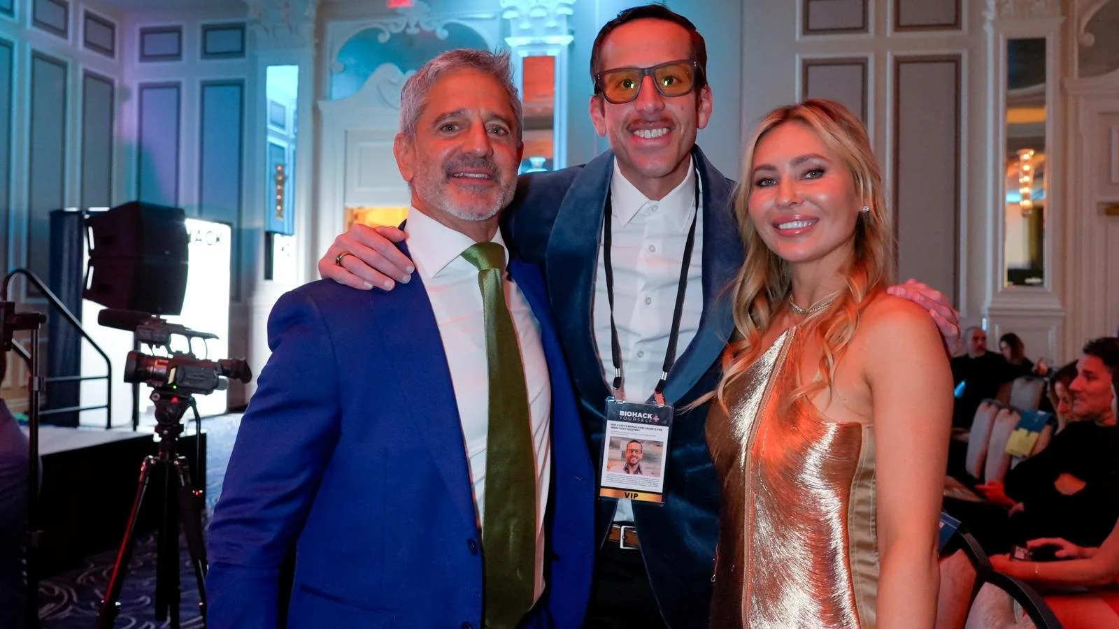Three people posing together at a conference, with a man in a blue suit and green tie on the left, a man with glasses and a conference badge in the middle, and a woman in a shiny gold dress on the right, in a banquet hall setting.