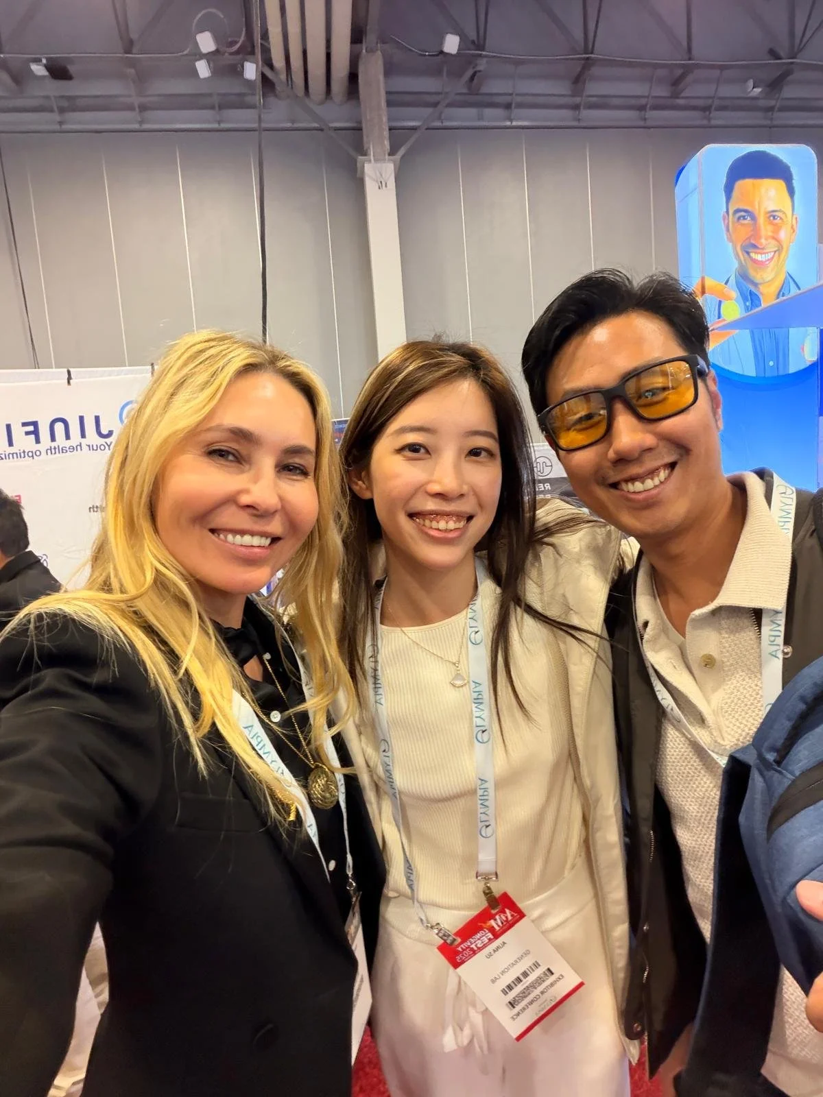 Three people smiling and posing for a photo at an indoor event, wearing conference badges with a large booth and a poster of a smiling man in the background.