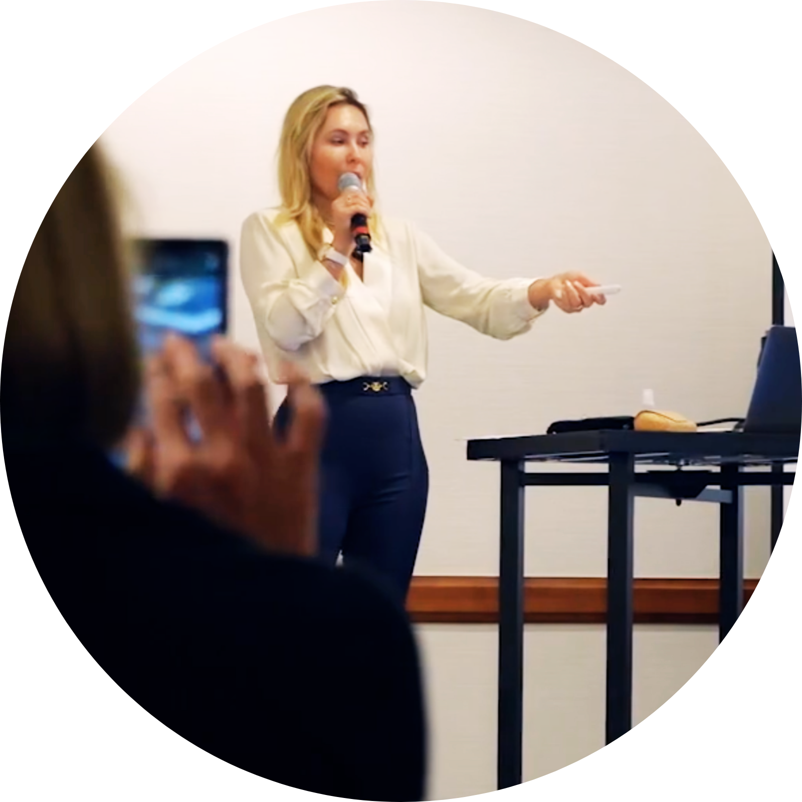 A woman with blonde hair wearing a cream blouse and dark pants is speaking into a microphone in a conference room.