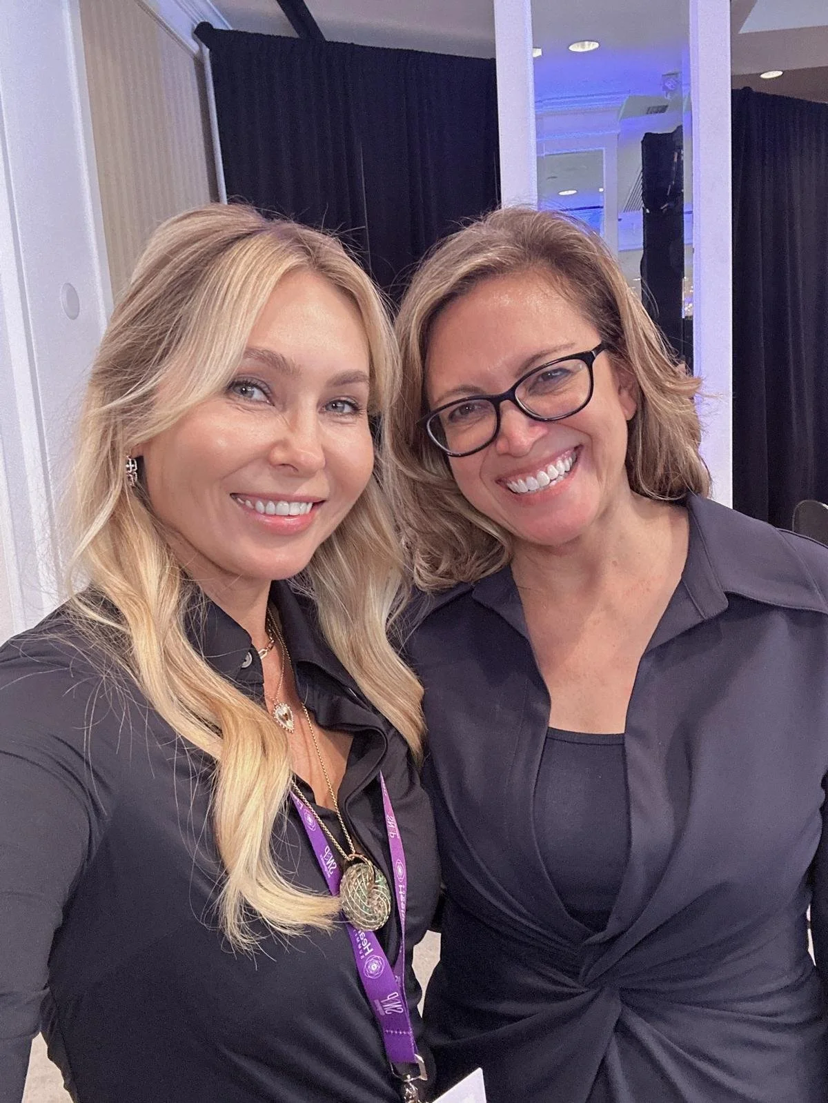 Two smiling women taking a selfie at an indoor event. One woman has long blonde hair and is wearing a black top with a purple lanyard and medals hanging from it. The other woman has short, wavy brown hair, glasses, and is also wearing a black top or 