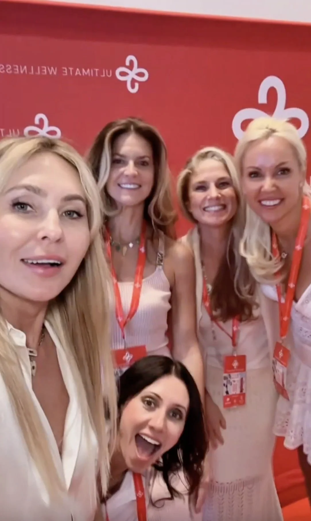 Group of five women smiling, taking a selfie, with a red background and white logo.