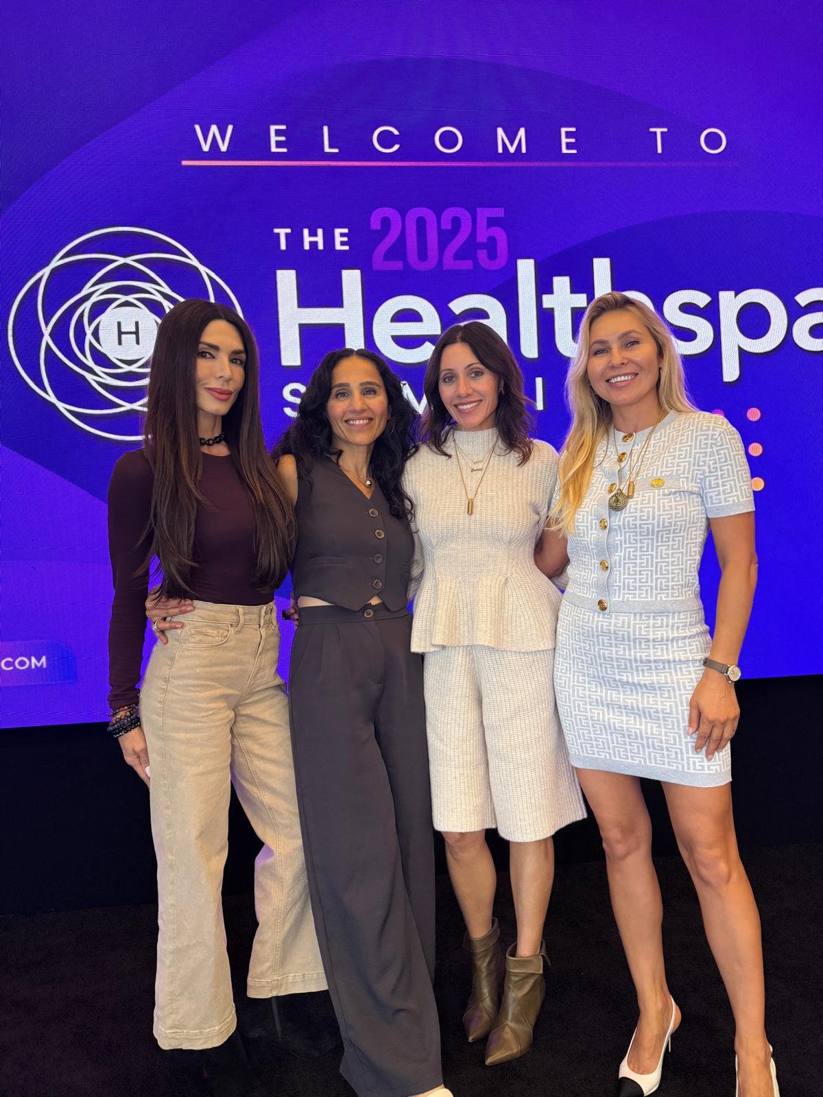 Four women posing in front of a large digital screen at the 2025 Healthspa event, smiling and dressed professionally.