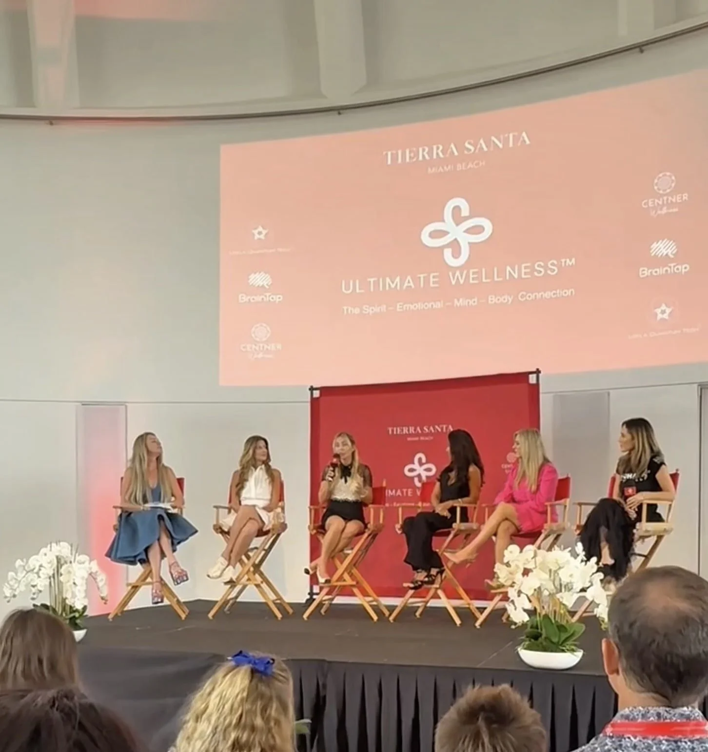 A panel of five women seated on stage during a wellness event, with a large screen displaying event details behind them and floral arrangements in front.