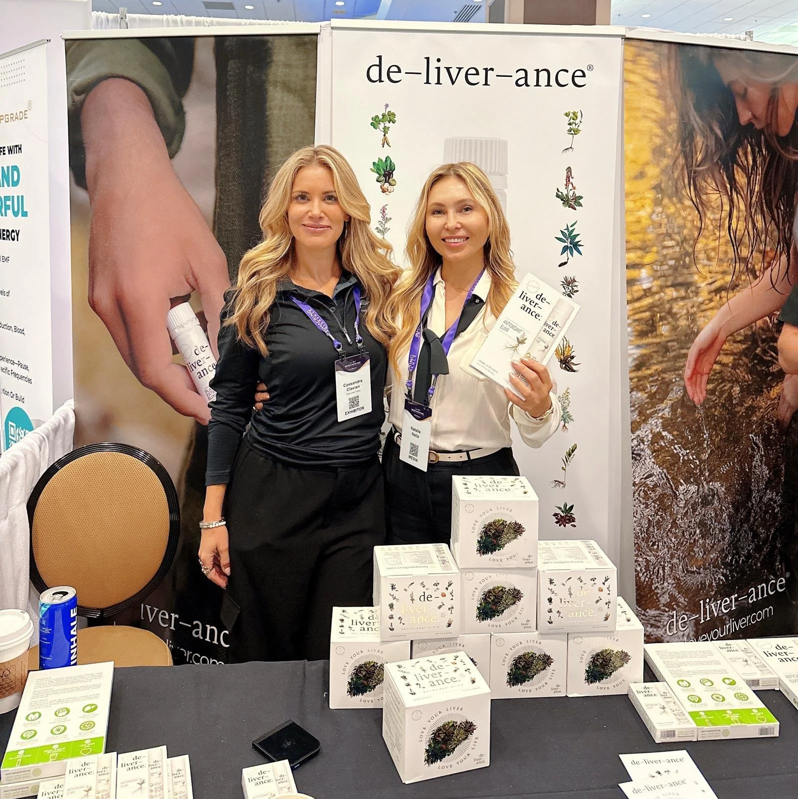 Two women standing behind a table displaying boxes of de-liver-ance herbal products at a trade show booth. They are smiling and holding brochures, with promotional banners in the background.