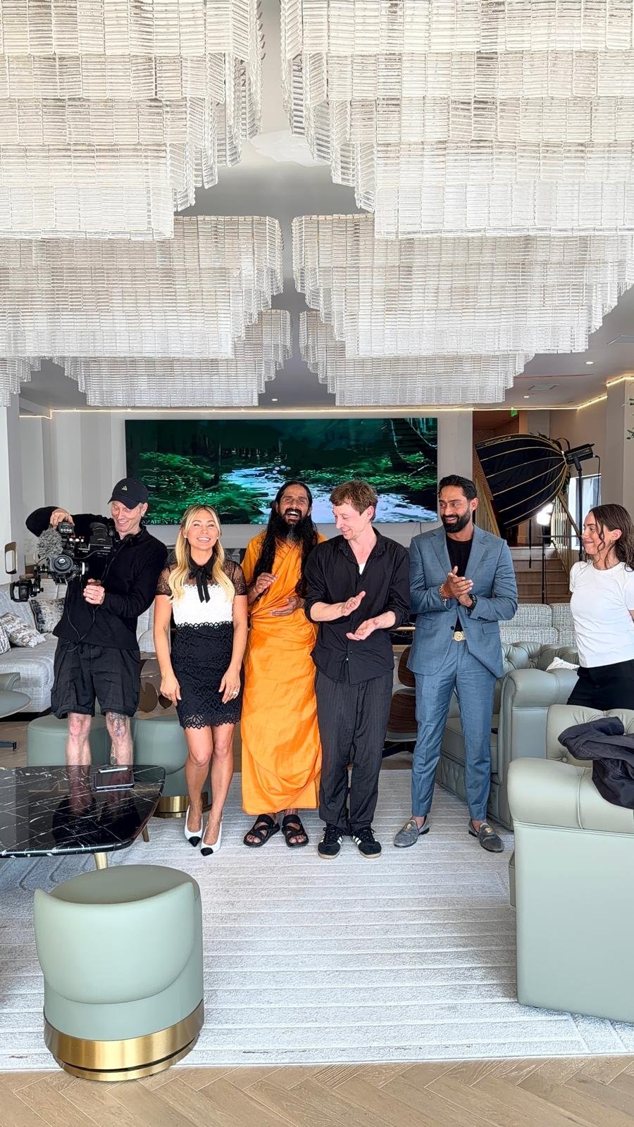 A group of six people standing in a modern living room, smiling and clapping, with a large screen displaying a nature scene in the background.