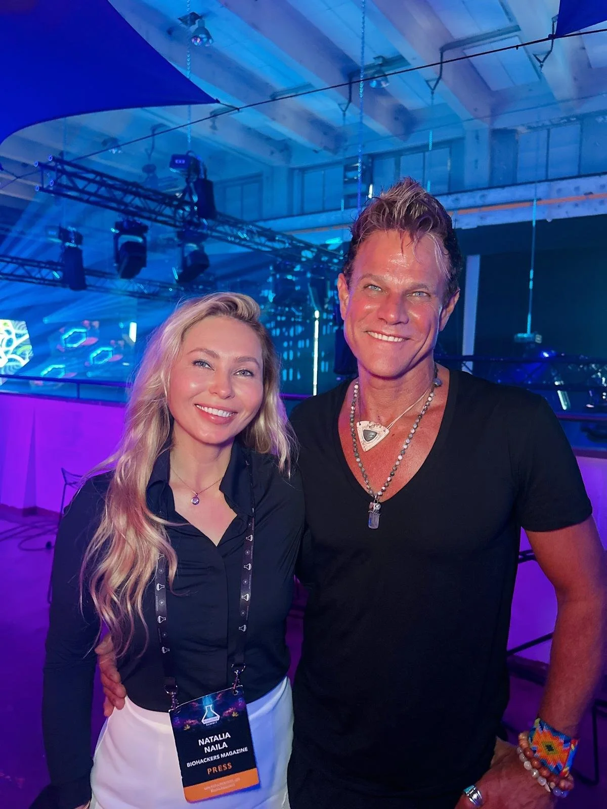 Two people smiling at an event with blue stage lighting, a woman wearing a black top and white skirt with a press badge, and a man in a black t-shirt wearing jewelry. The background shows a venue with lighting equipment and a wooden ceiling.