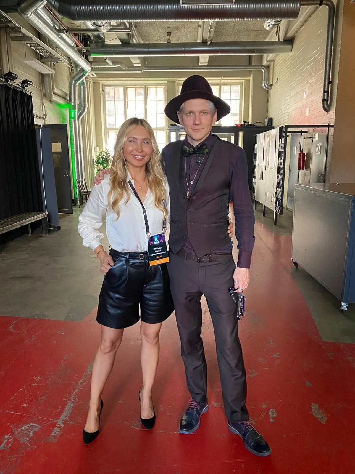 A woman and a man are standing together indoors, smiling at the camera. The woman has long blonde hair and is wearing a white blouse, black leather shorts, and black high heels. The man, taller than the woman, is wearing a black hat, dark vest, dark 