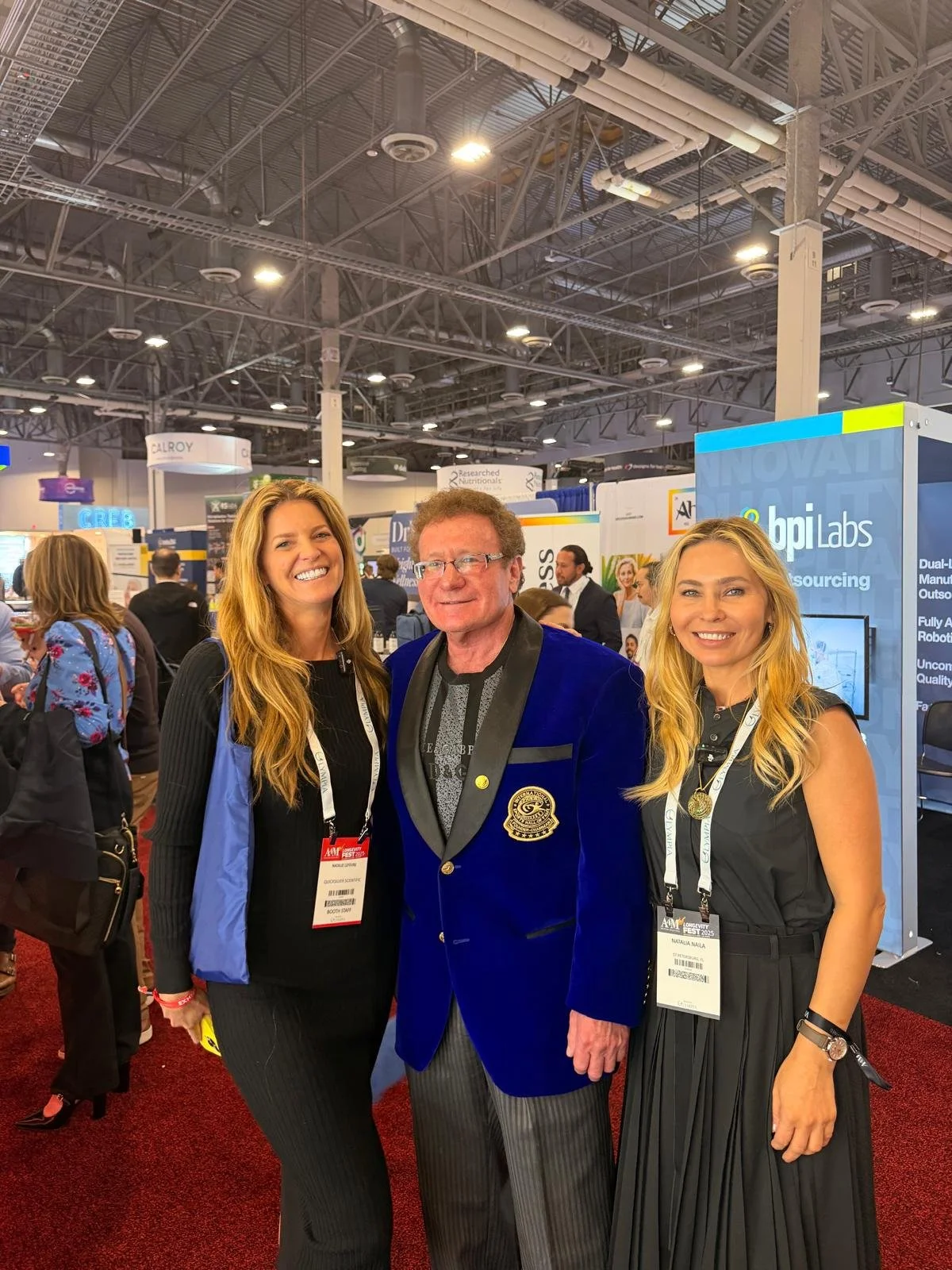 Three people smiling at a conference, two women and a man in a blue blazer with a badge, standing in front of exhibition booths.