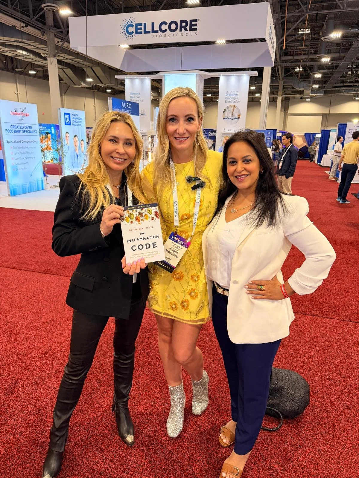 Three women posing together at a conference, one holding a book titled 'The Inflammation Code', with exhibition booths and signs in the background.