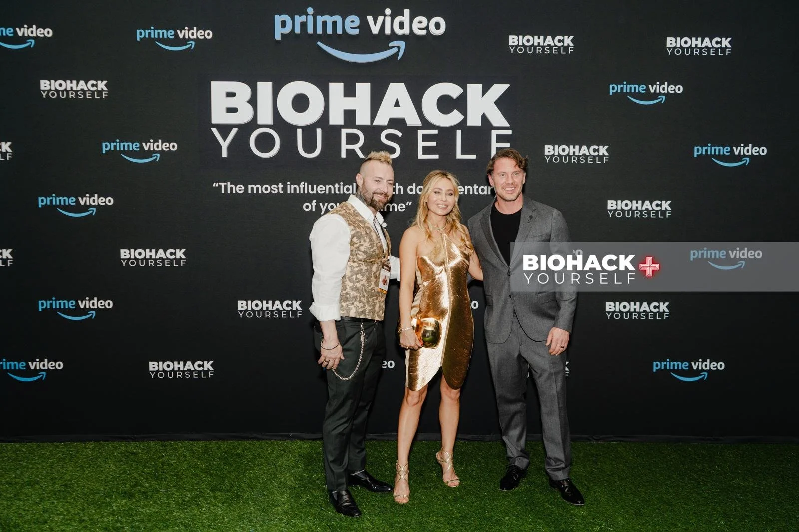 Three people, two men and one woman, standing together on grass in front of a large black backdrop with the words 'Biohack Yourself' and logos for Prime Video and Biohack Yourself, smiling at the camera.
