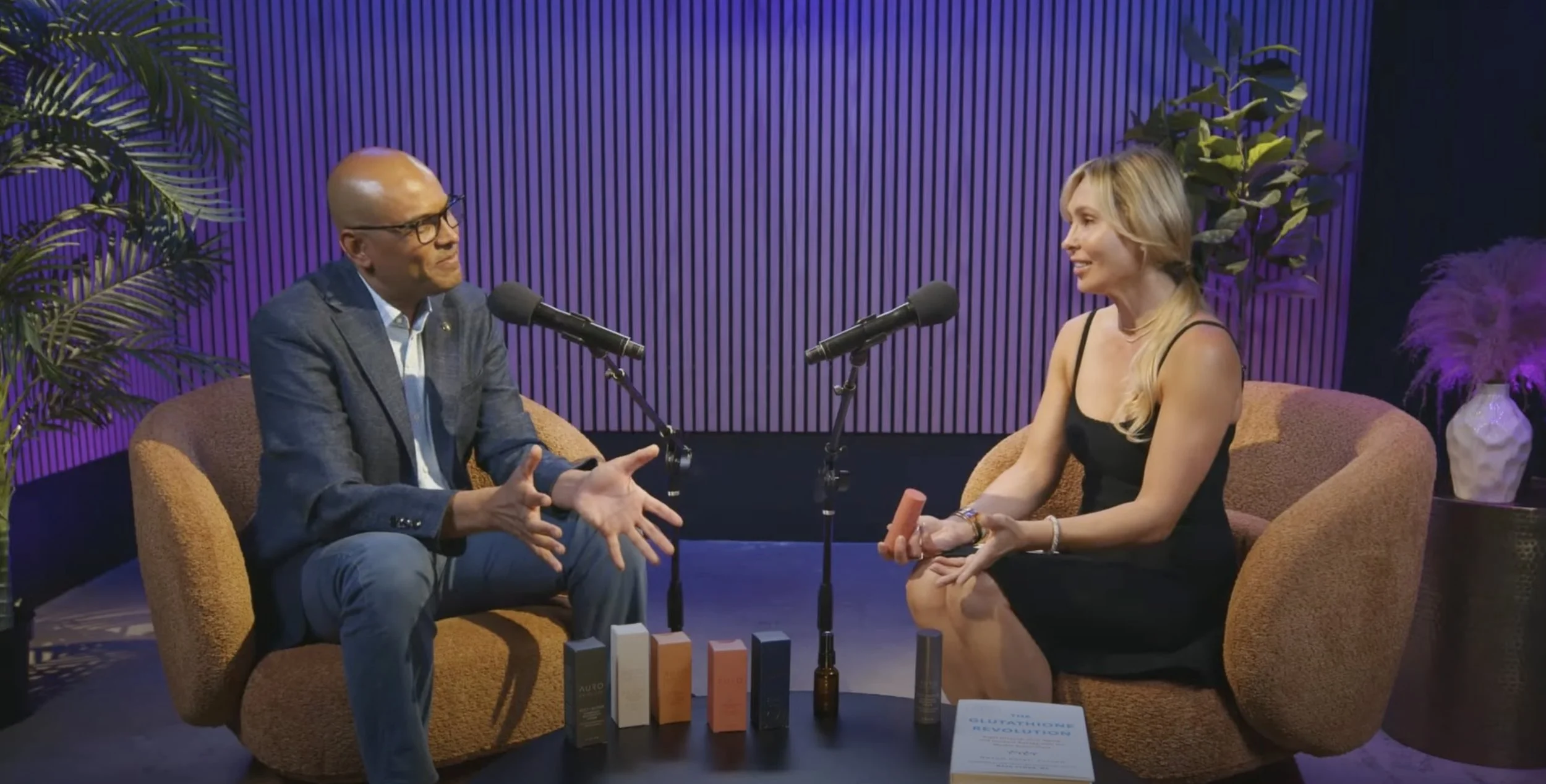 A man and a woman sit on brown couches in a recording studio with purple and black walls, engaged in a conversation with microphones in front of them. There are various skincare or cosmetic products on a table between them, along with a book titled 'GLOW UP' on beauty.