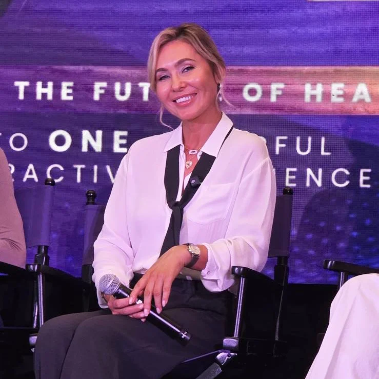 A woman with short blonde hair, wearing a white blouse and black skirt, sitting on a chair and holding a microphone, smiling at a conference or panel discussion.