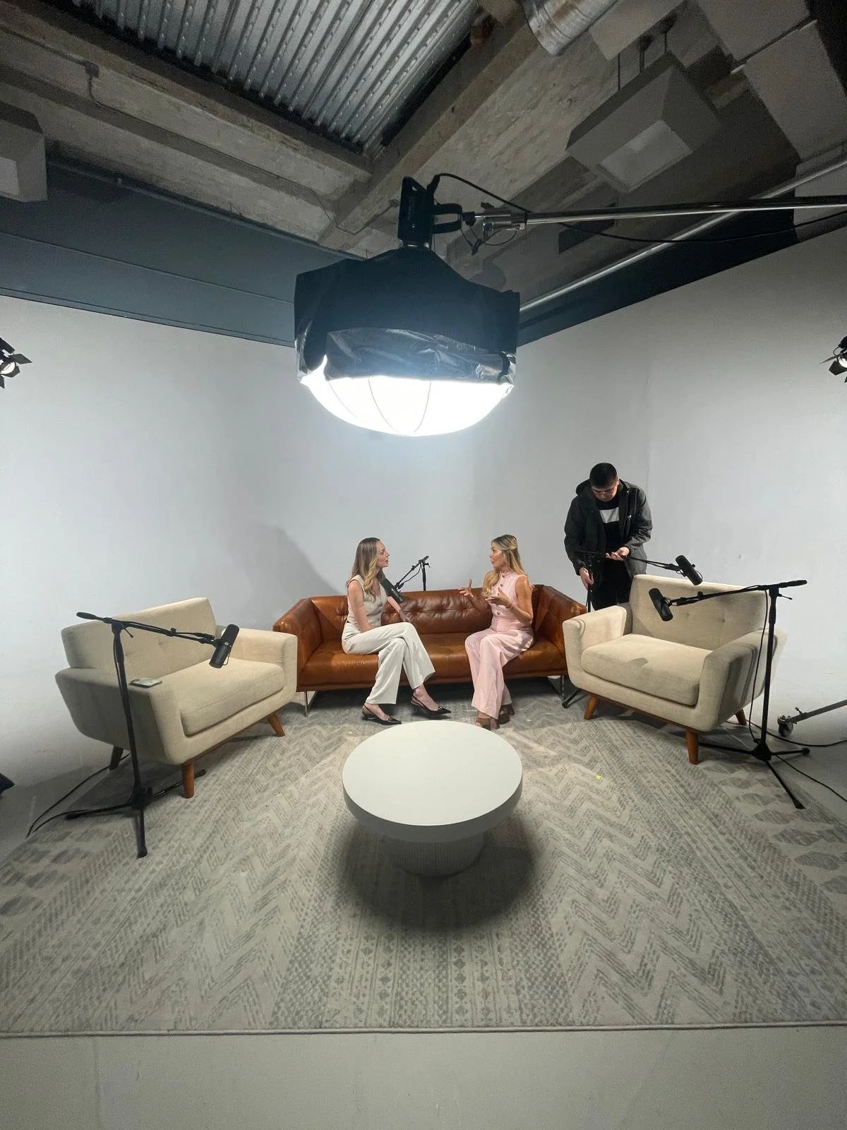 Two women sitting on a brown leather sofa, engaged in conversation, with a man standing nearby behind beige sofas with microphones, in a white-walled recording studio with professional lighting.