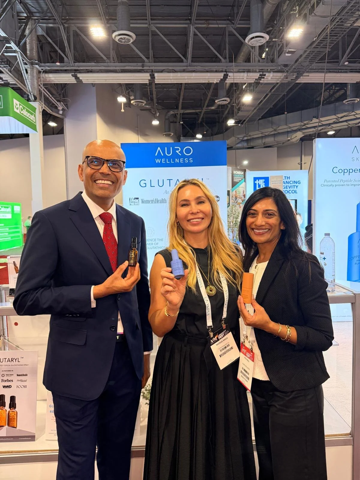 Three people at a wellness booth holding skincare or cosmetic products and smiling at the camera