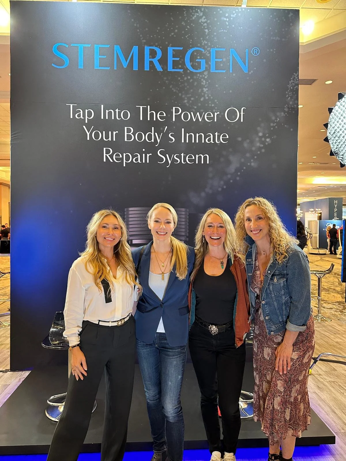 Four women standing in front of a large, dark blue promotional display board that reads 'STEMREGEN - Tap Into The Power Of Your Body's Innate Repair System' at a conference or event.