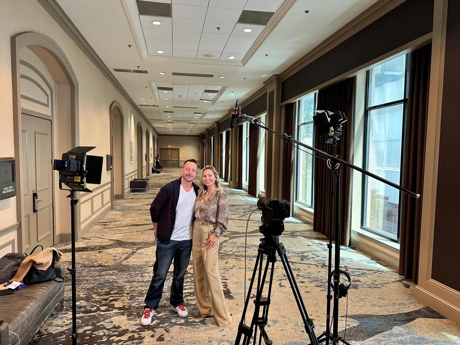 Two people, a man and a woman, stand together in a well-lit hallway with large windows, next to camera equipment used for filming or photography. The man wears a dark jacket, white shirt, and jeans, and the woman wears a patterned blouse and beige pa