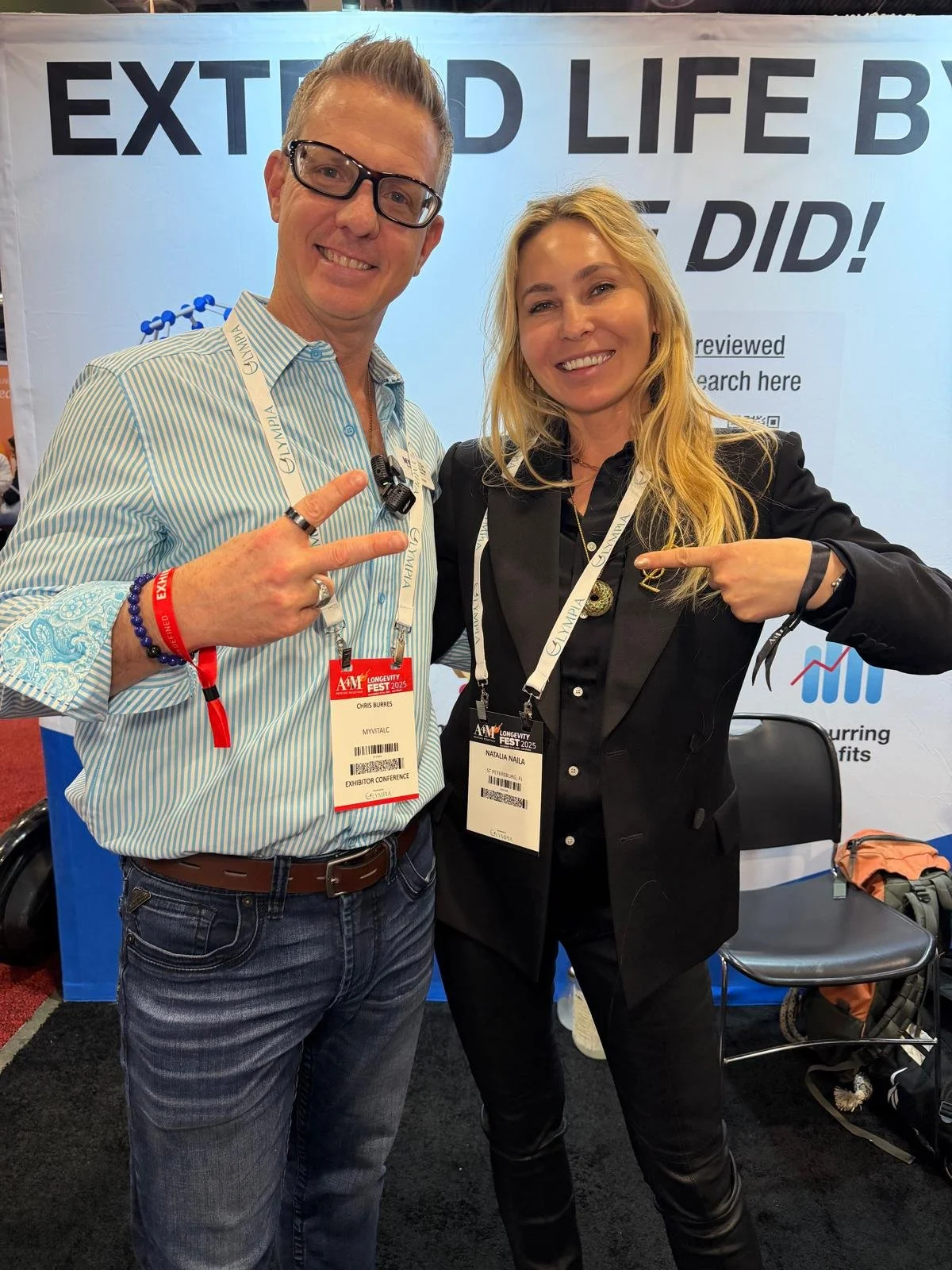 Two smiling people, a man and a woman, posing at a conference booth. The man is wearing glasses, a striped shirt, and has a conference badge that says 'Chr Burres.' The woman has long blonde hair, is dressed in black, and has a badge that reads 'Nata