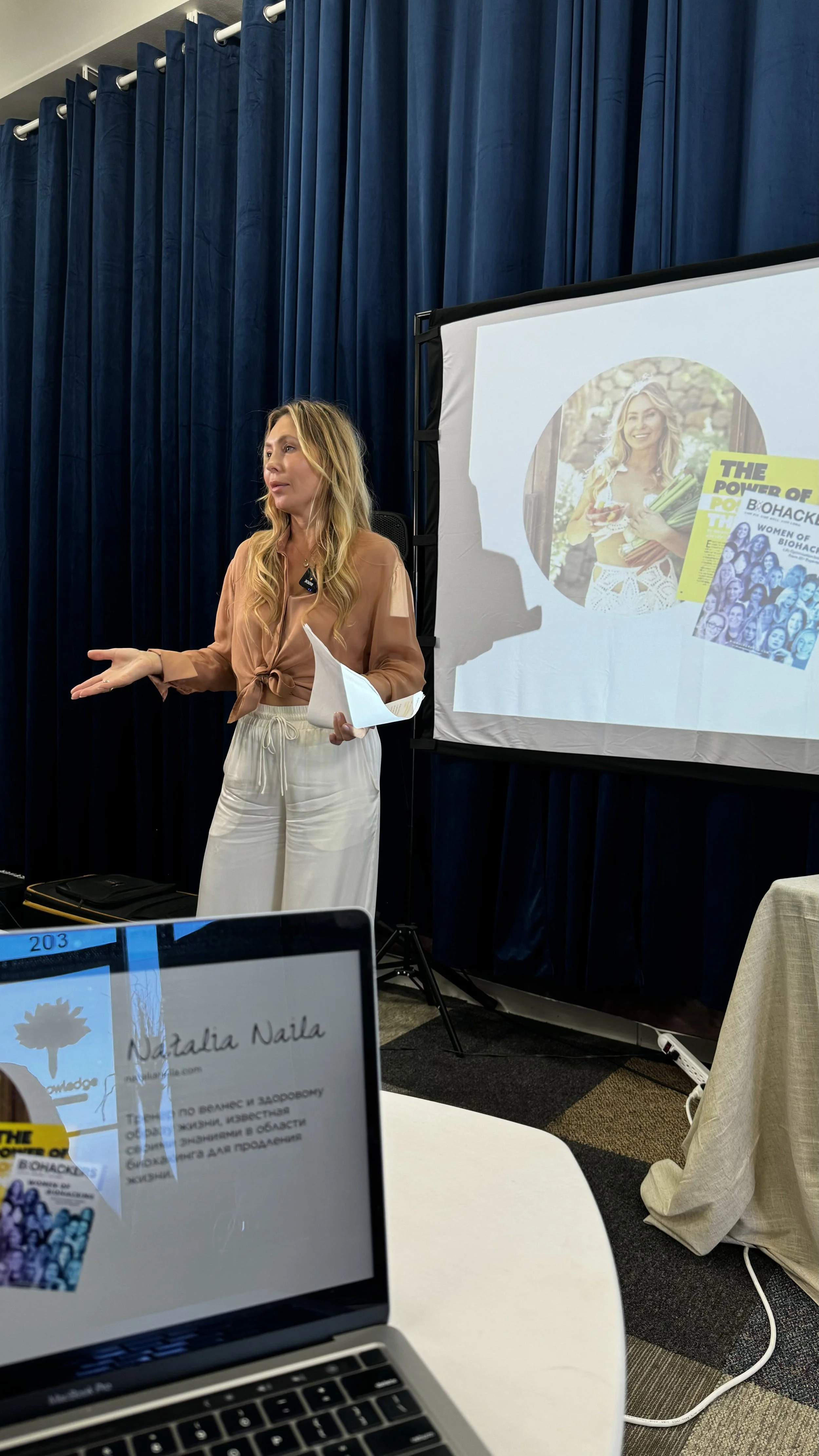 woman giving a presentation in front of a screen with a colorful image of a woman holding vegetables and a magazine, with blue curtains in the background.