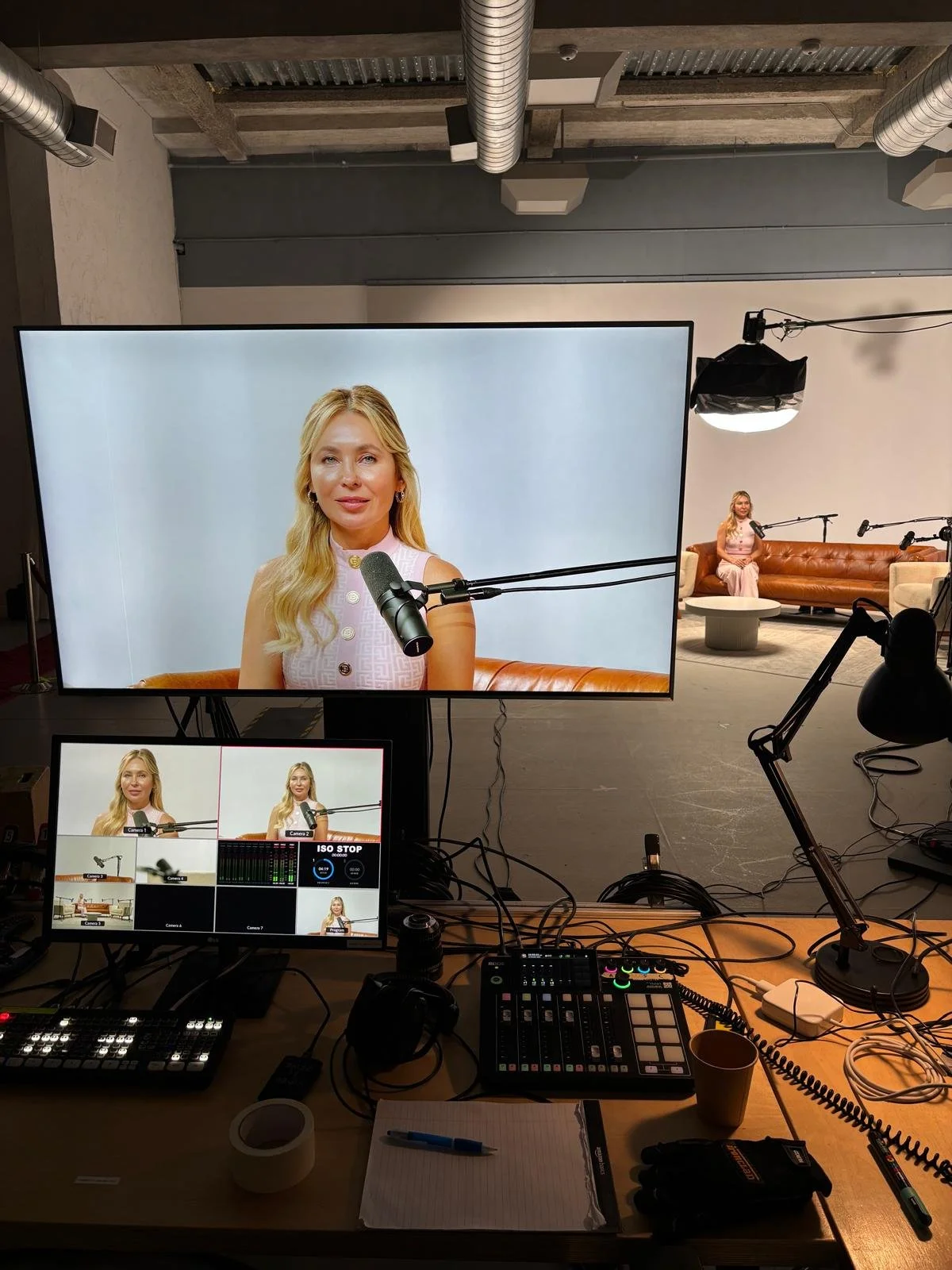 Video shoot setup with a large screen displaying a woman speaking into a microphone, studio lighting, a second woman on a sofa, and video control equipment on a table.