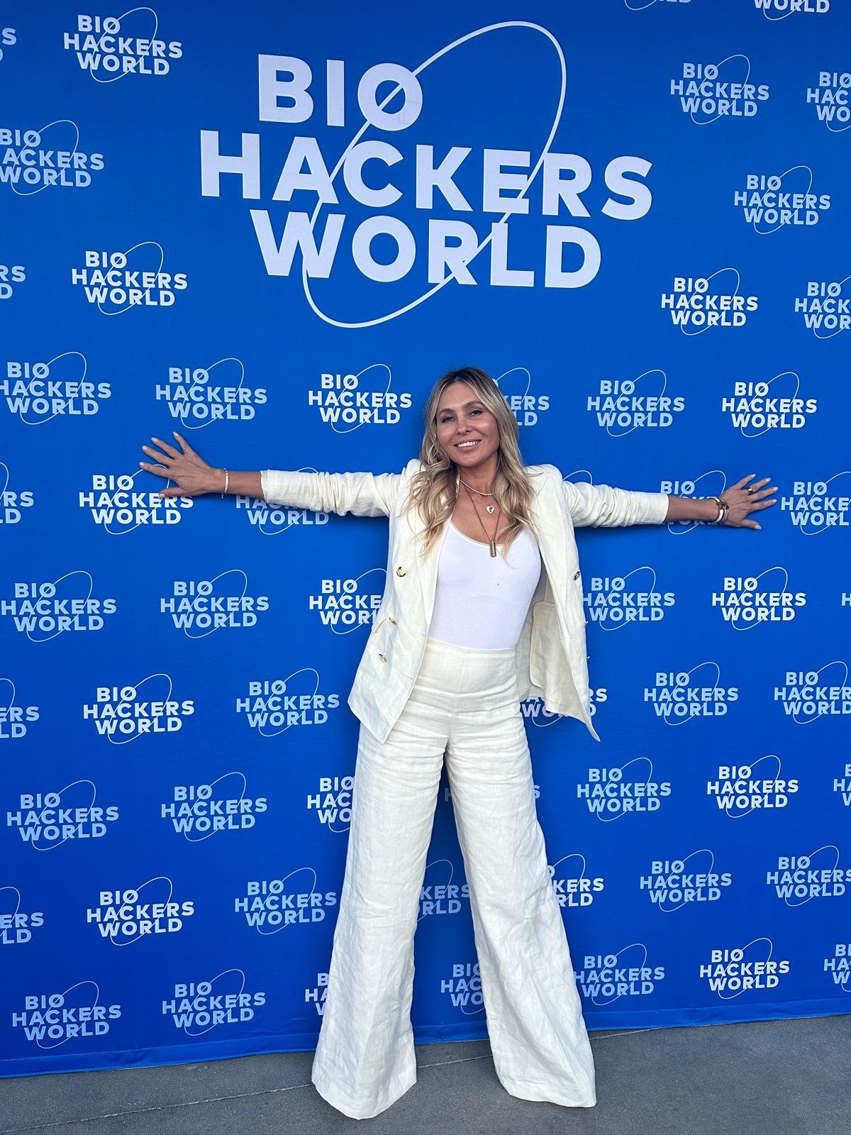 A woman in a white suit with a white top, standing with arms outstretched in front of a blue backdrop with multiple 'Bio Hackers World' logos.