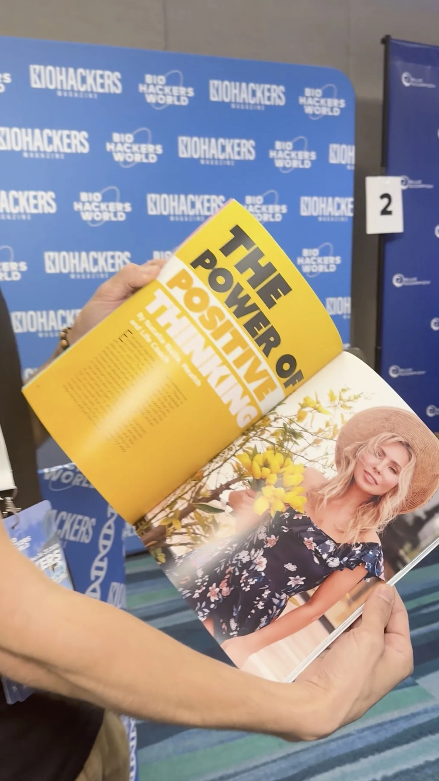 A person holding an open magazine with a yellow headline page that says "The Power of Positive Thinking" and an interior photo of a woman with blonde hair wearing a black floral dress and a wide-brim straw hat, standing outdoors with yellow flowers.