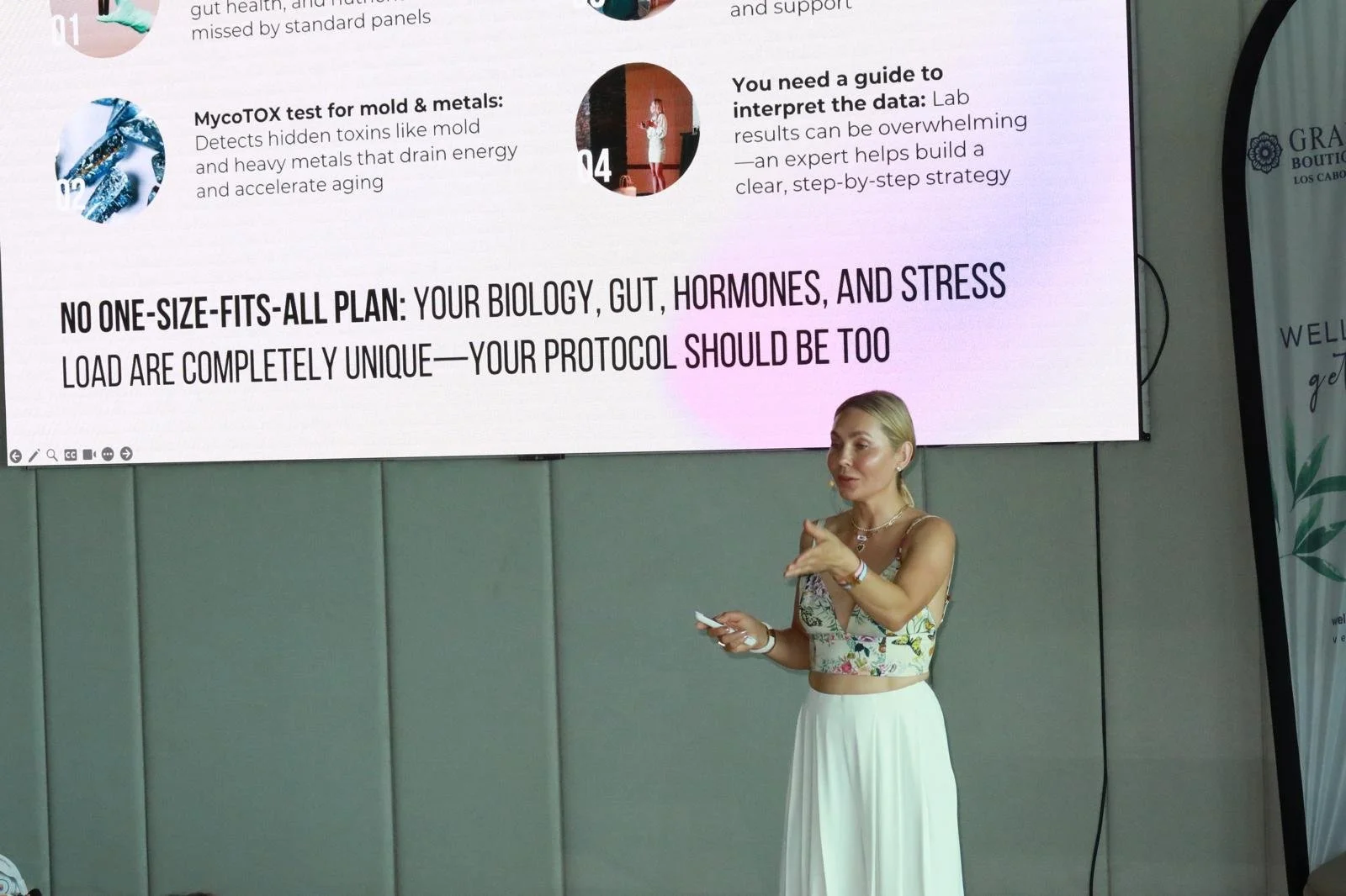 A woman giving a presentation in front of a large screen displaying text about biology, gut, hormones, stress, and lab testing.