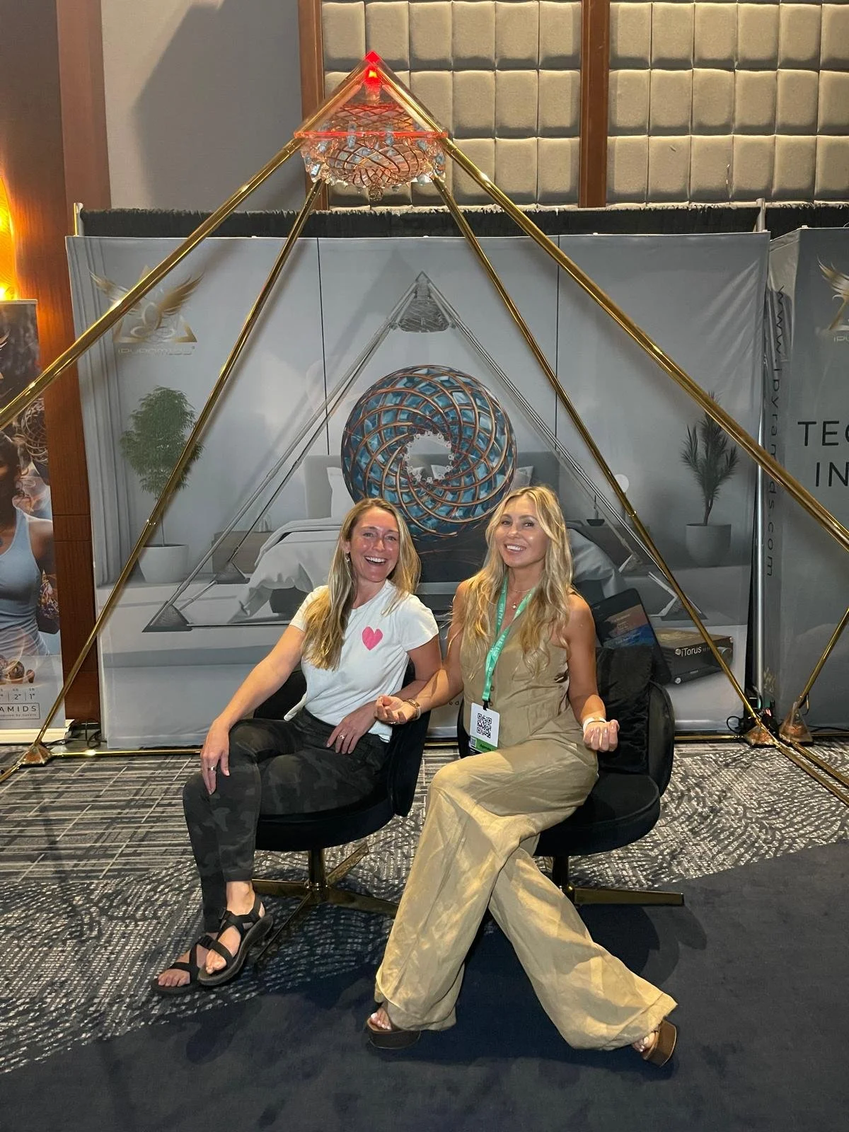Two women sitting in chairs at a convention with a geometric tent structure overhead. They are smiling and holding hands, with a large decorative spherical sculpture and a bed display in the background.