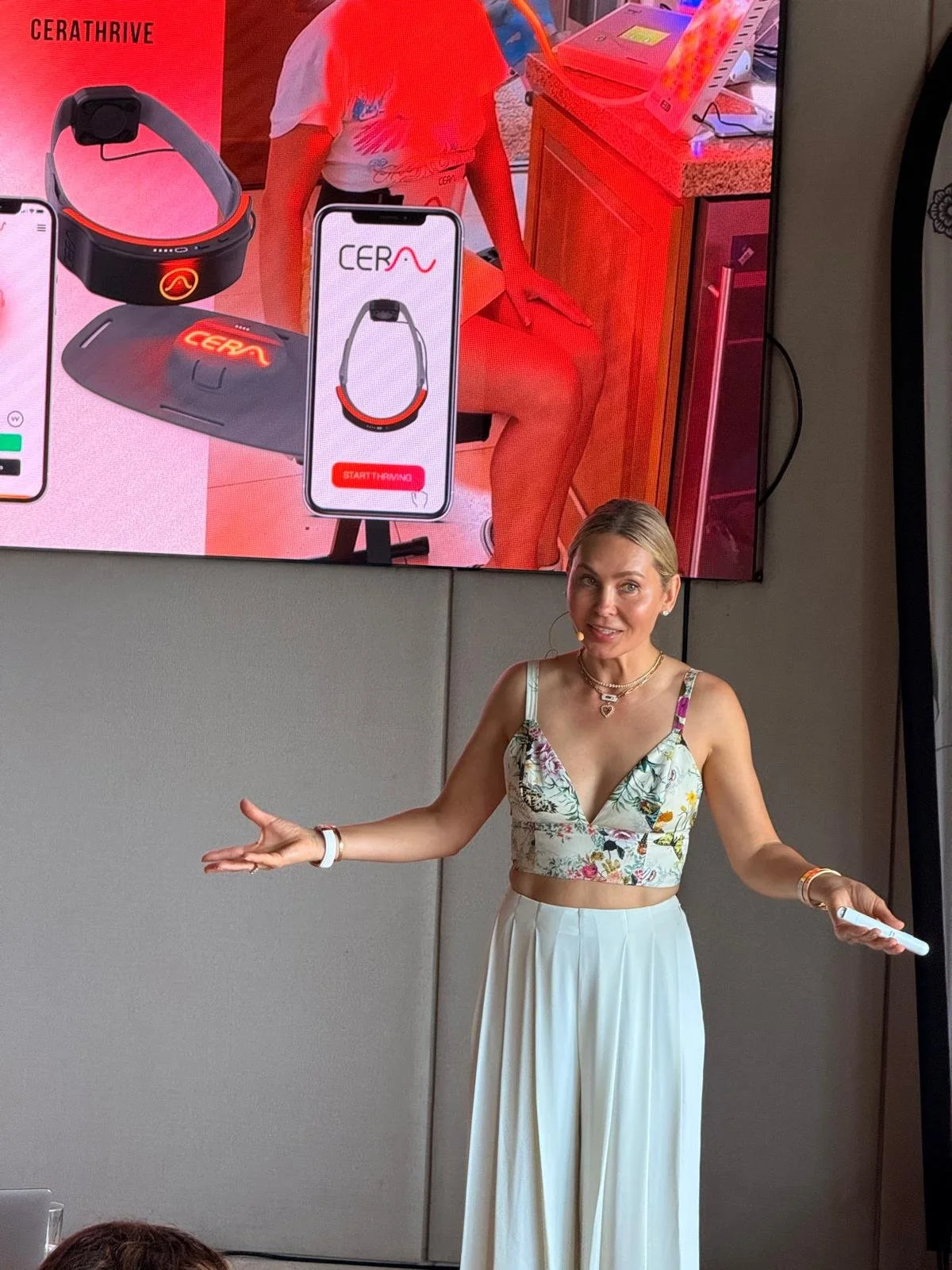 A woman in a floral crop top and white wide-leg pants pointing with both hands in front of a large screen displaying a health and fitness device called CERATHRIVE, including a headband and a mobile app interface.