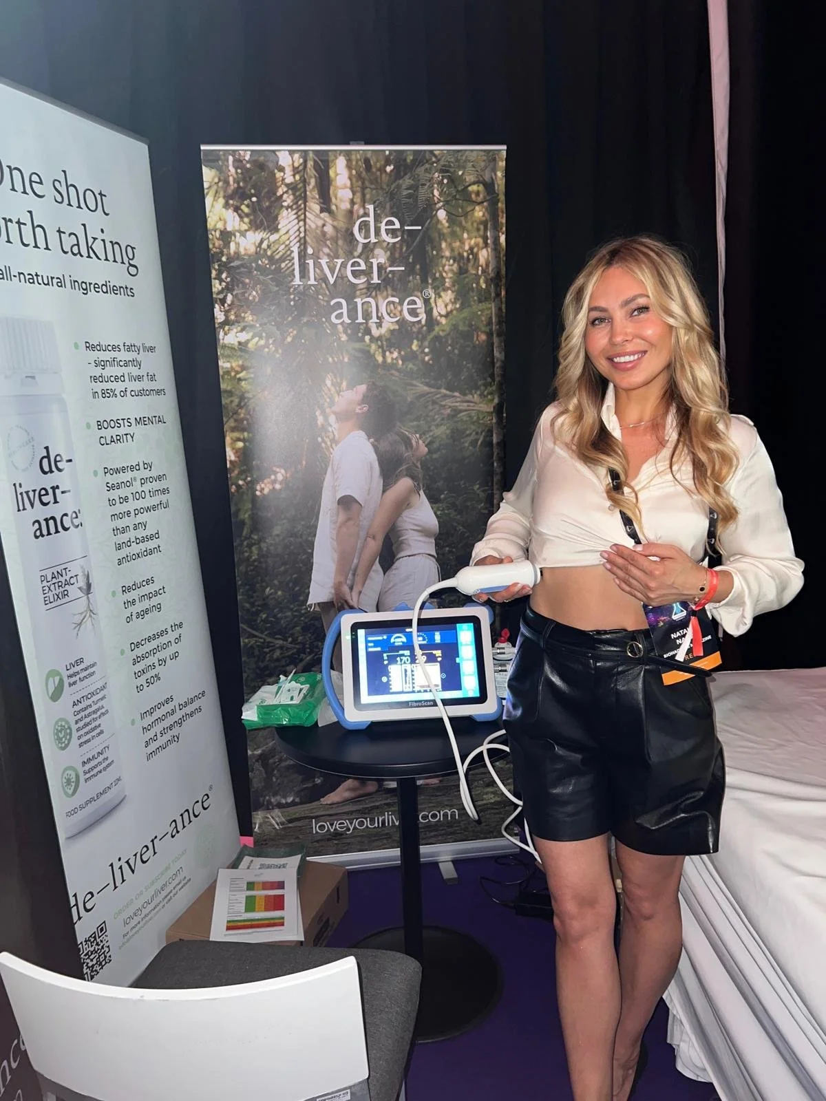 A woman smiling at a booth with health-related promotional displays and a device used for a medical scan or therapy, with a large banner behind her showing an outdoor scene with people in nature.