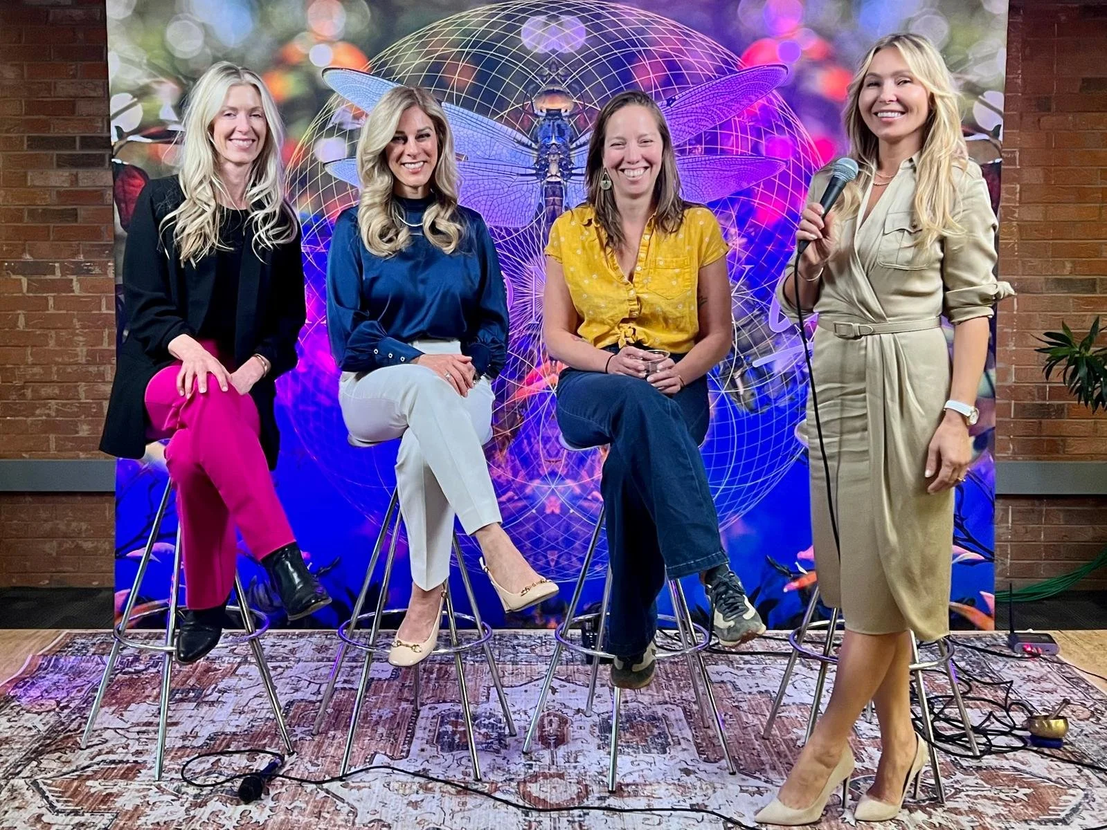 Group of four women sitting on high stools in front of a colorful, digital-themed backdrop, with one woman standing and holding a microphone.
