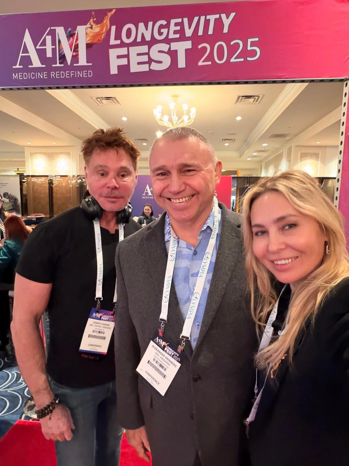 Three people standing under a large purple and pink sign that reads "A4M Longevity Fest 2025," smiling. They are wearing conference lanyards with name tags, in a well-lit conference hall.