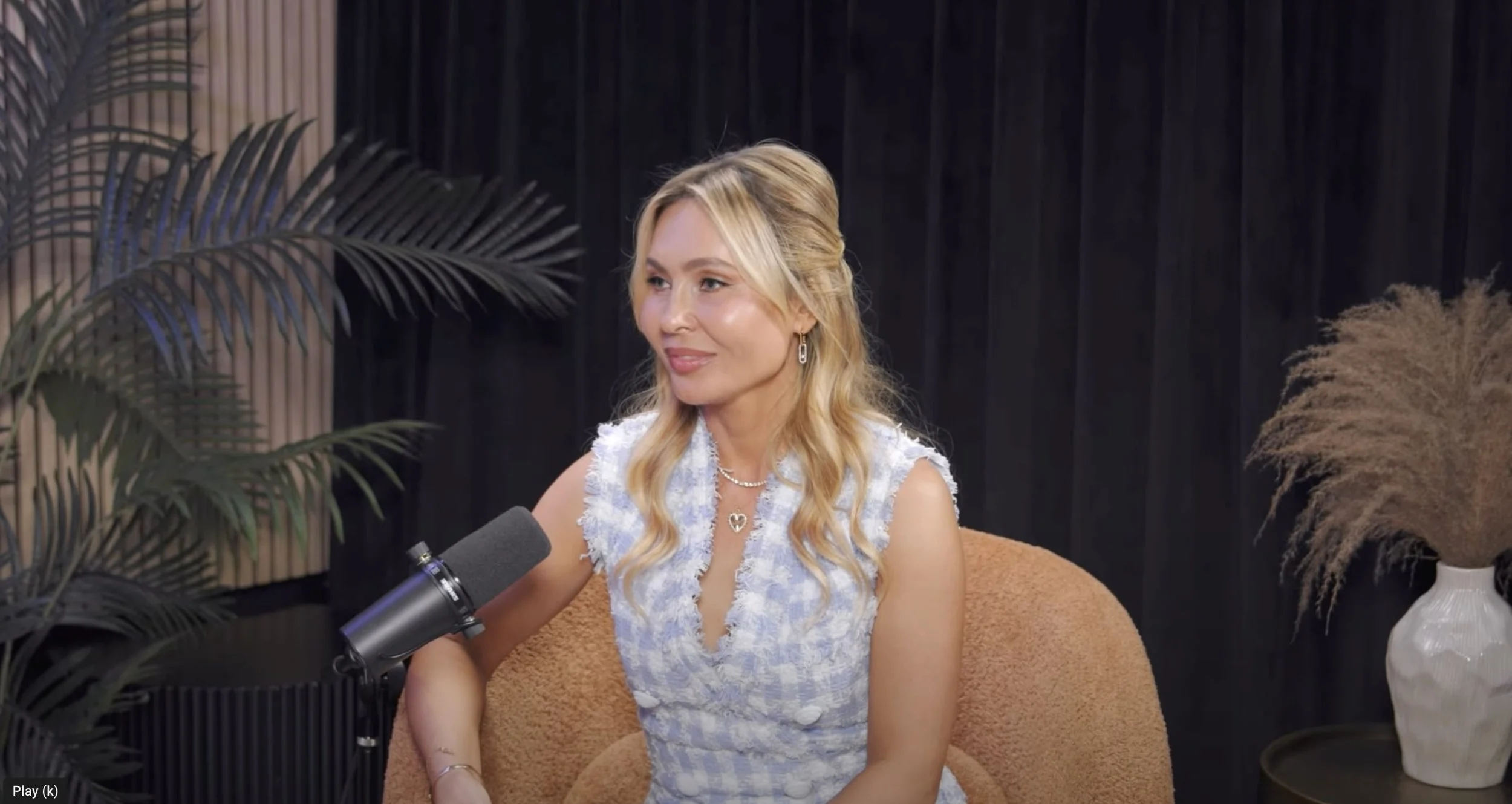 A woman with long blonde wavy hair sitting on a brown armchair, smiling, during an interview with a microphone in front of her. The background features black curtains, decorative plants, and dried pampas grass in a white vase.