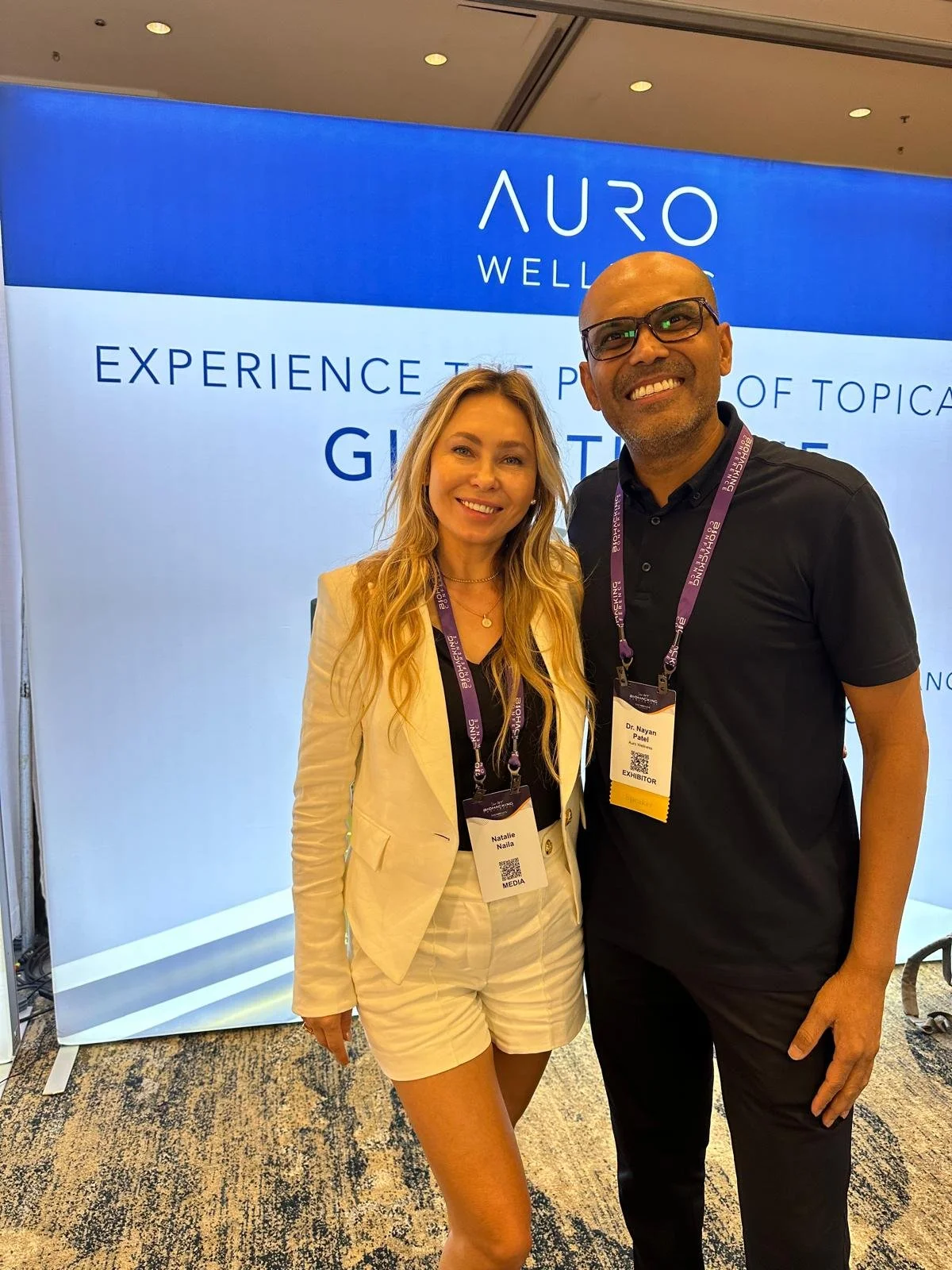 Two people standing in front of a blue and white display at a conference or expo, smiling, with name tags around their necks.