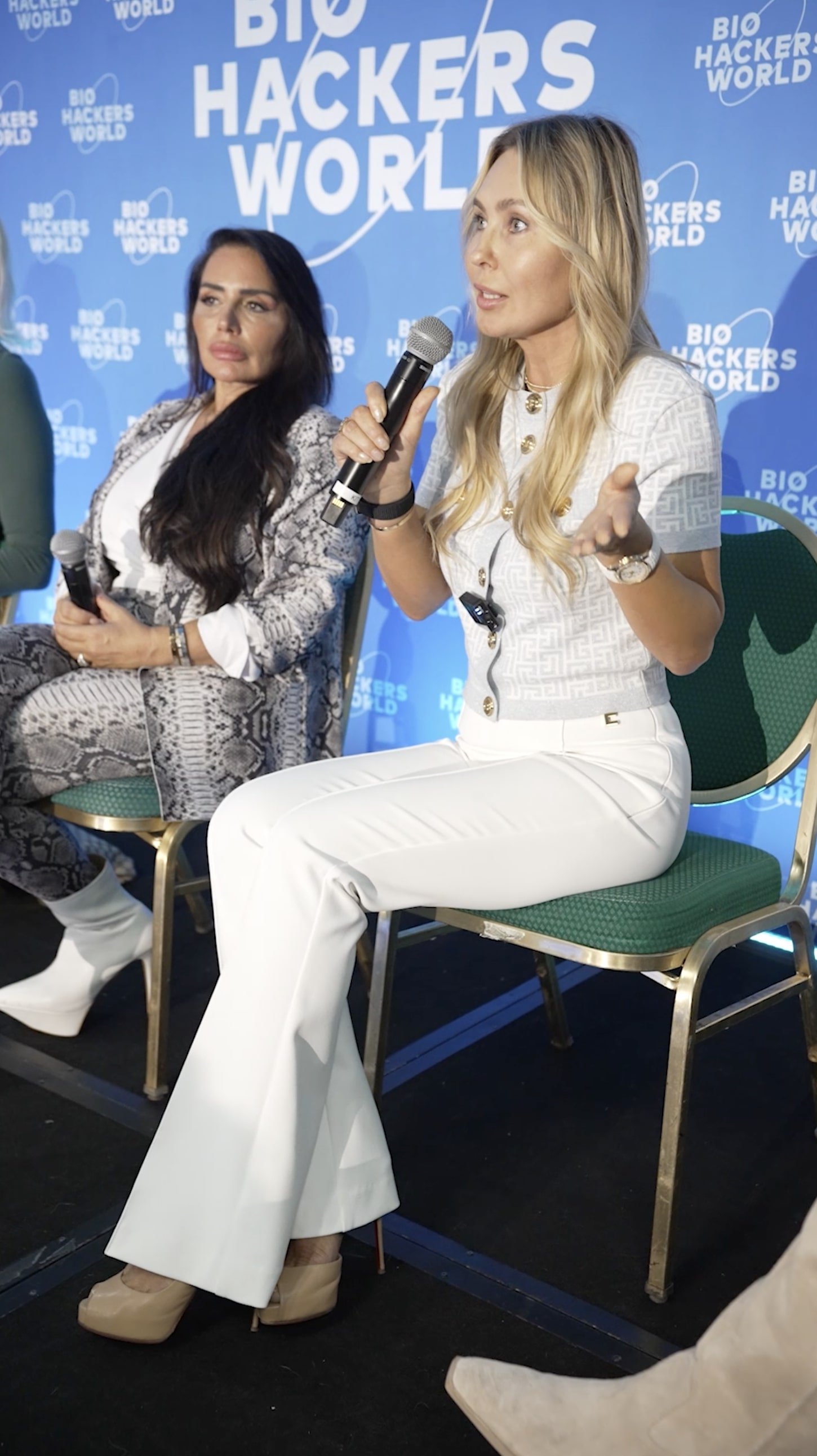 Two women seated on stage at BioHackers World conference, one in white pants and the other in snakeskin-patterned suit, both holding microphones, with a blue conference backdrop.